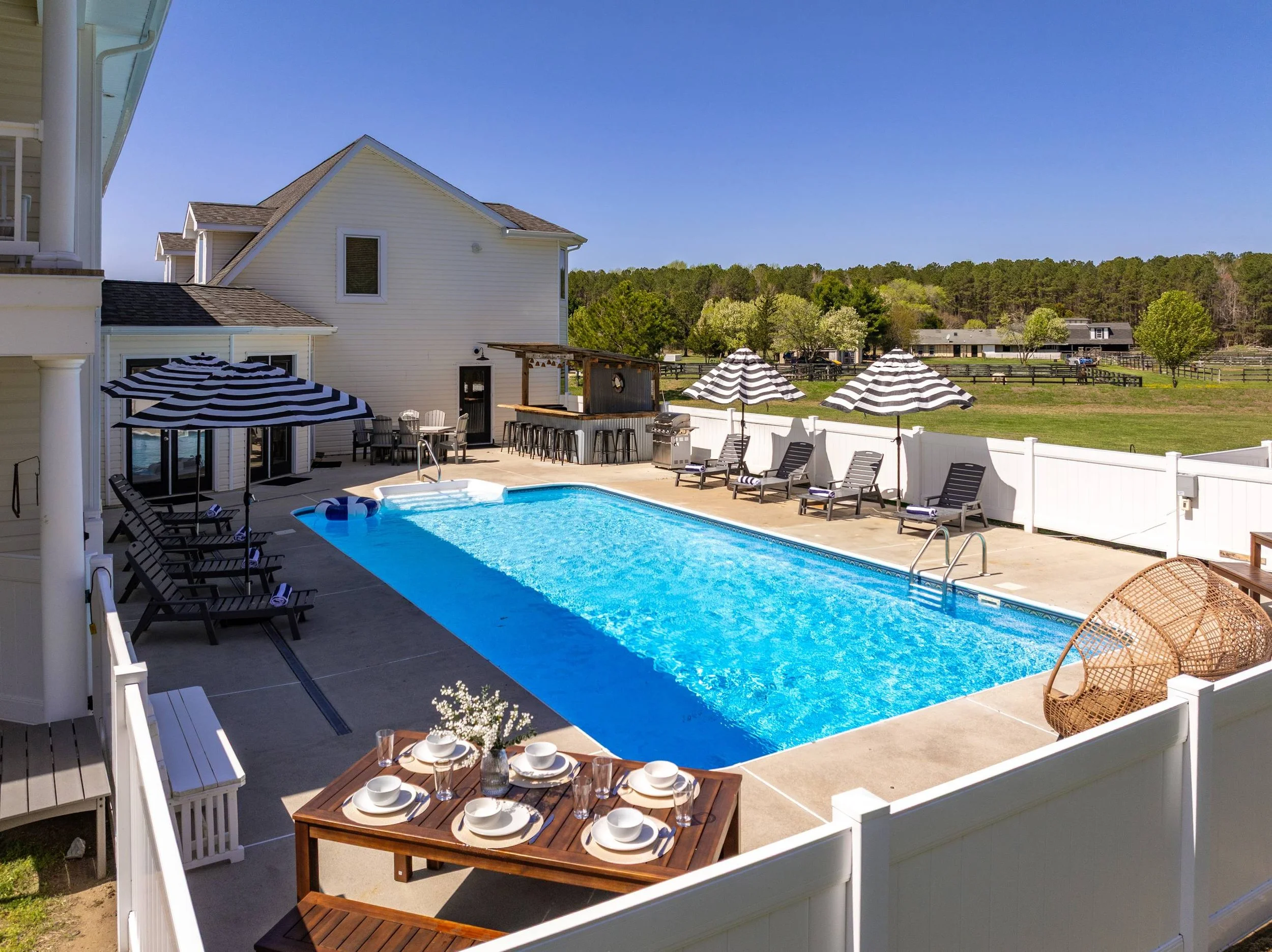 A backyard swimming pool area with lounge chairs, striped umbrellas, a dining table set with plates and glasses, surrounded by a white fence. There are houses and trees in the background under a clear blue sky.