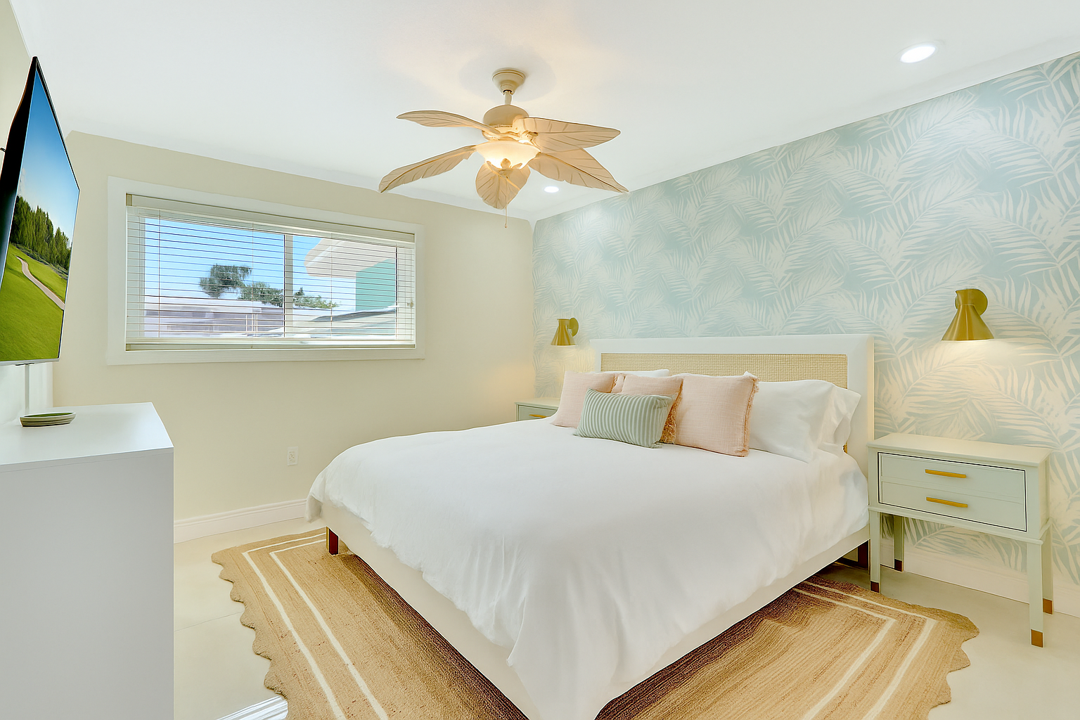 Bright bedroom with a white bed, pastel pillows, a beige woven headboard, nightstands with gold lamps, a ceiling fan with leaf-shaped blades, a window with blinds, and a tropical wallpapered accent wall.