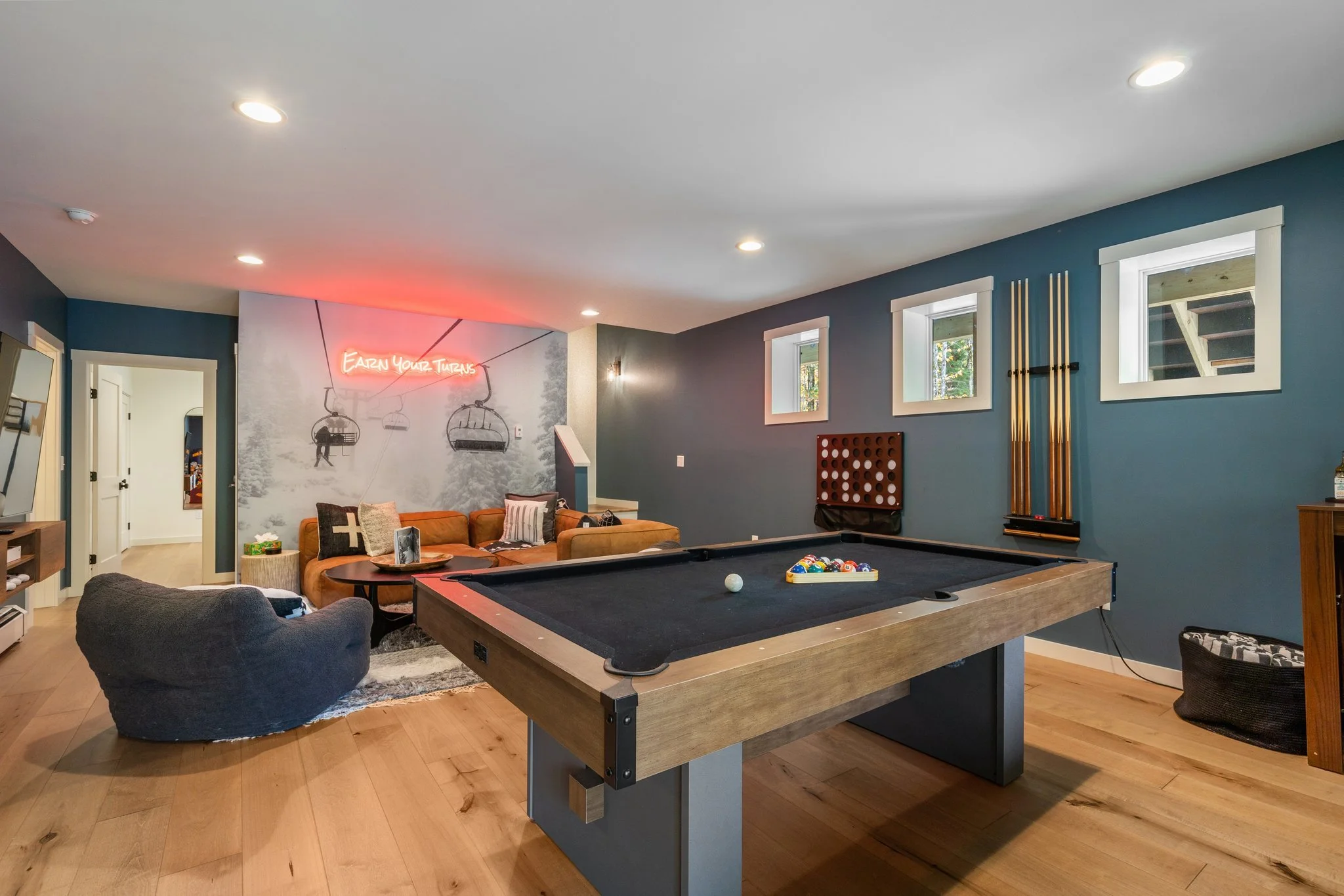 Living room with a pool table, sofa, armchair, and a stone side table. Walls are dark teal with three small windows, a neon sign reading 'Earn Your Turns', and a wall art with pool cues.