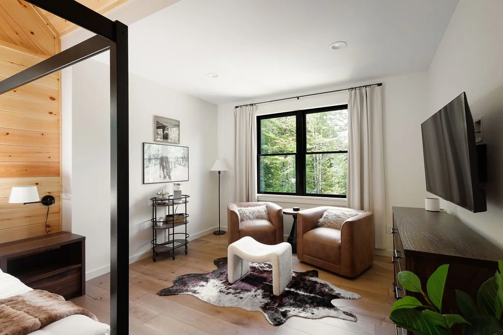 Living room with two tan armchairs, a cowhide rug, a window with white curtains, a wall-mounted TV, a black side table, a white floor lamp, and a wooden cabinet.