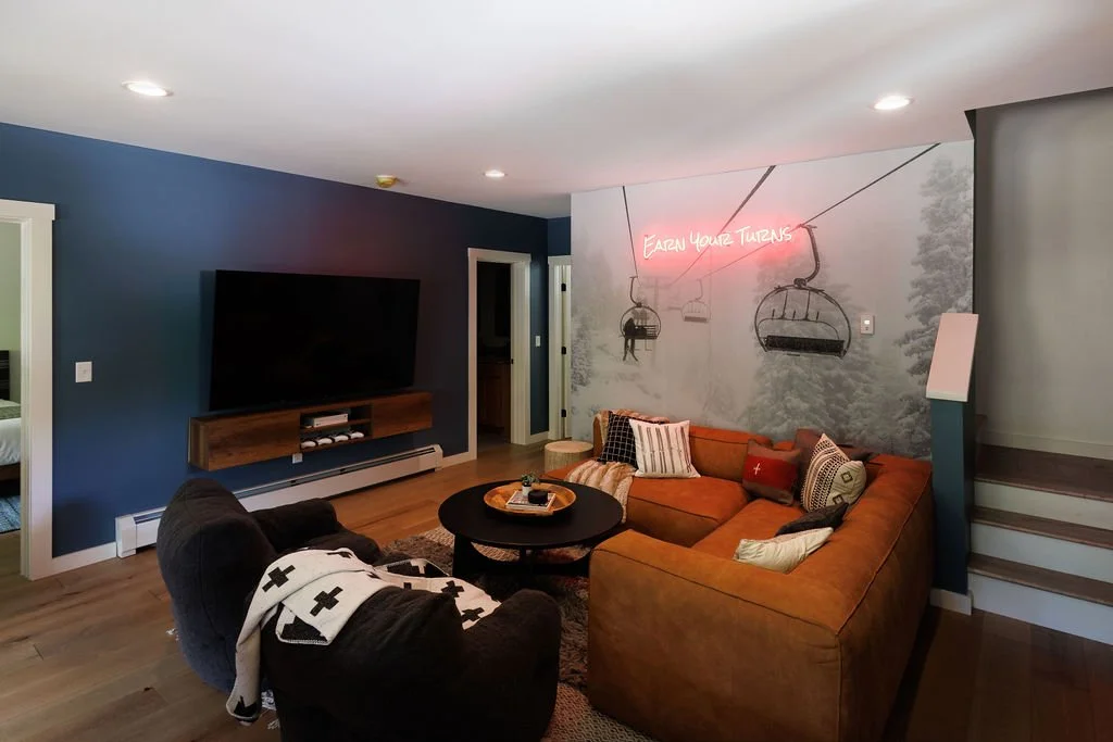 Living room with an orange sectional sofa, black armchair, wall-mounted flat-screen TV, wood cabinet with electronic devices, and a neon sign that says 'Earn Your Turns' above mounted hanging chair illustrations on the wall.