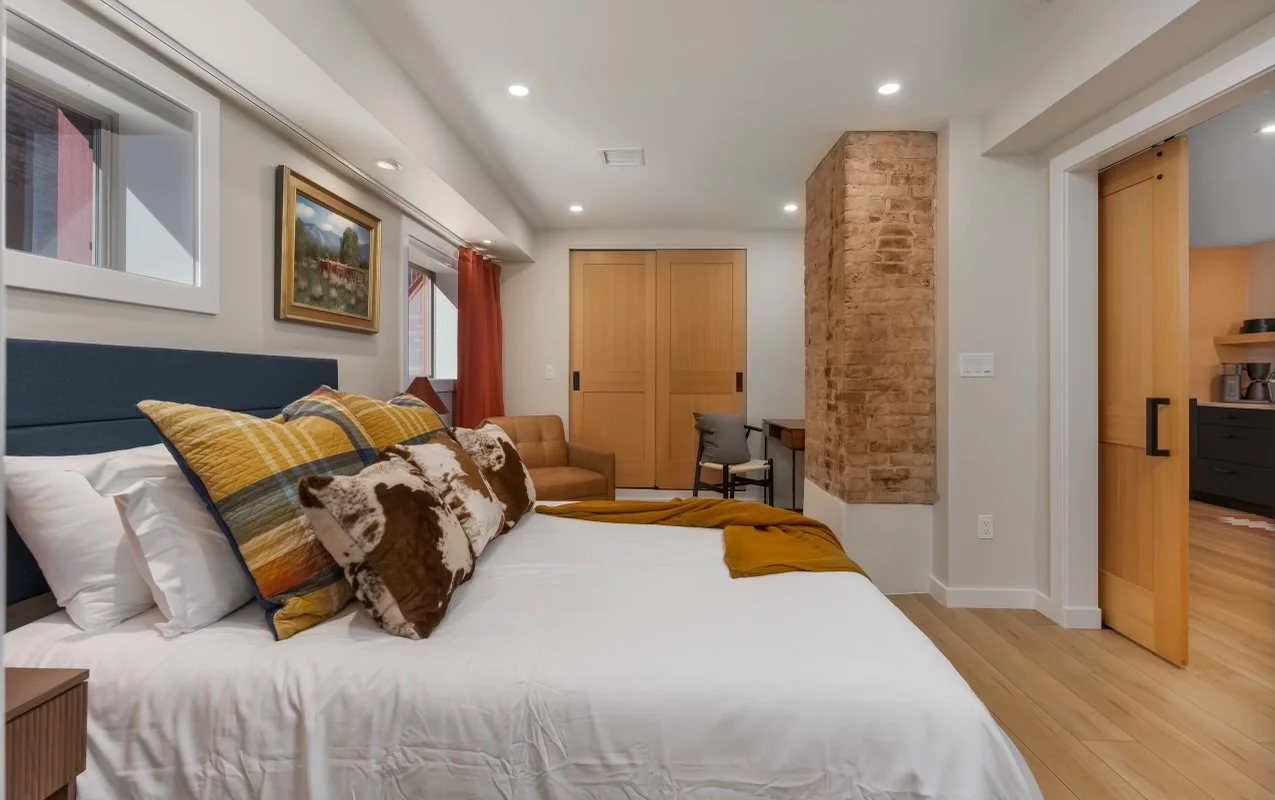Bedroom with a bed, decorated with multiple pillows, a window with red curtains, and a brick wall section.