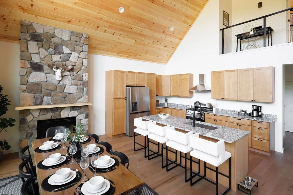 Open-concept kitchen and dining area with a stone fireplace, wooden ceiling, and modern decor, showcasing a dining table set for six and a kitchen with wooden cabinets, granite countertops, and a row of white barstools.