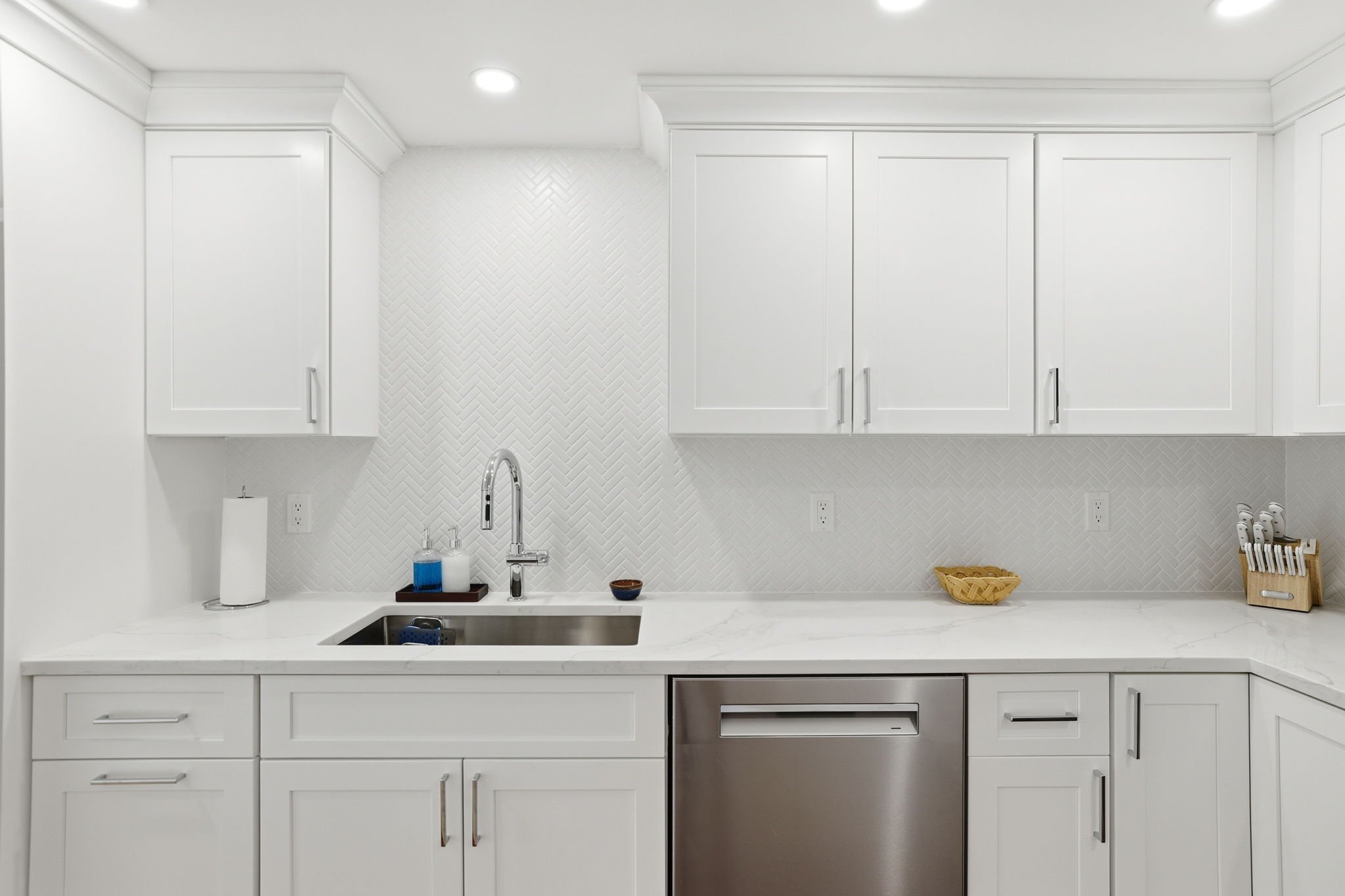 White kitchen with white cabinets, marble countertop, stainless steel dishwasher, and a white backsplash with a herringbone pattern. Coffee supplies and a small basket are on the counter.