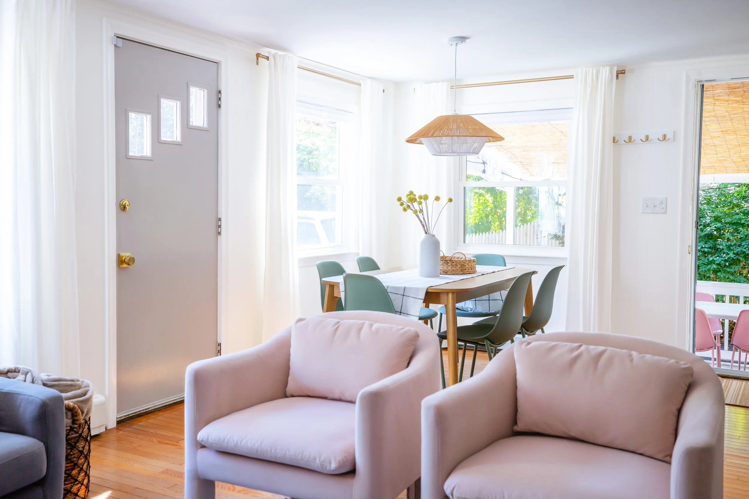 Bright dining area with windowed doors and white curtains, a table with a white tablecloth, a large white vase with yellow flowers, surrounding beige upholstered chairs, and a bamboo pendant light.