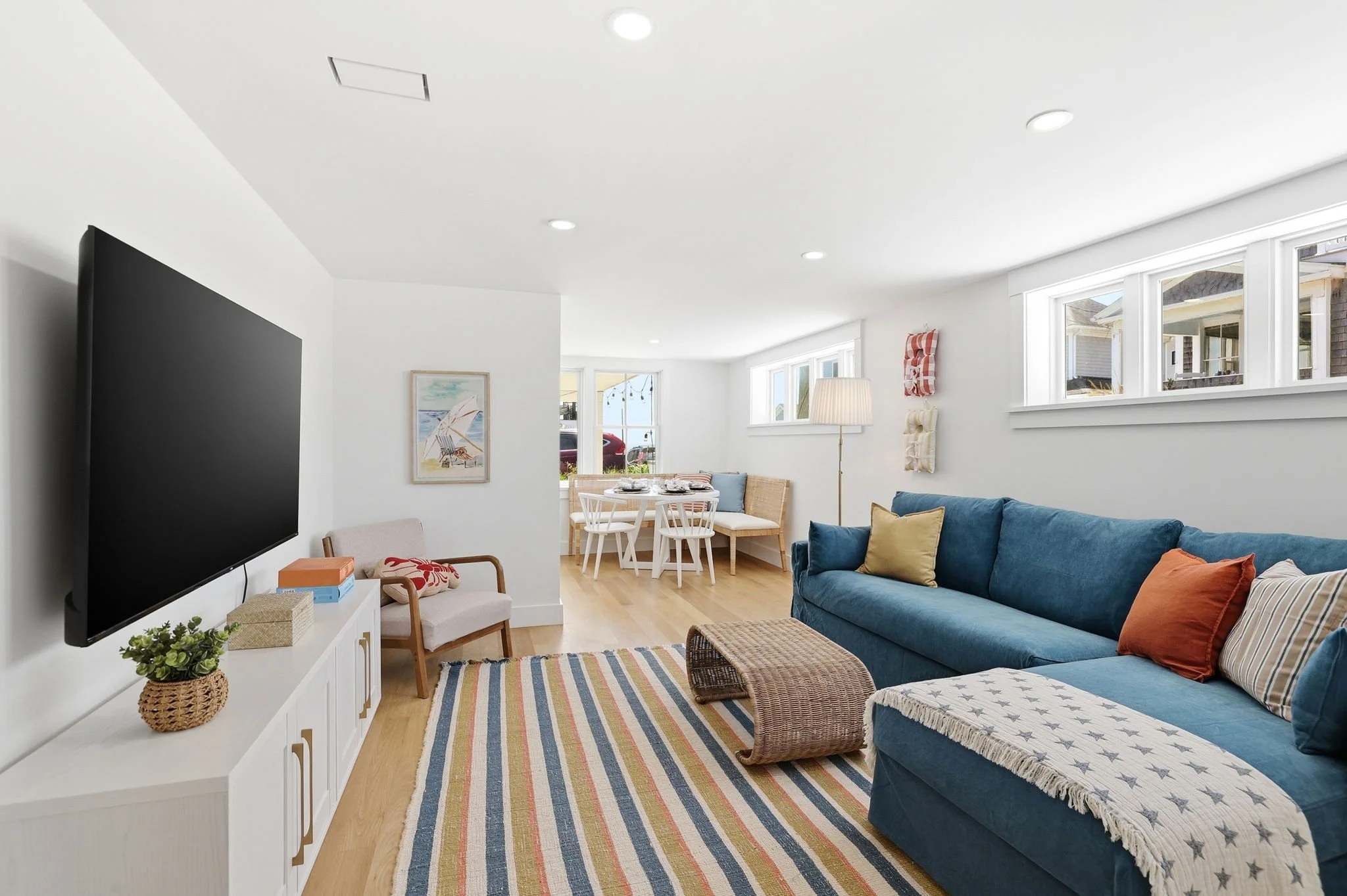 Living room with white walls, large windows, a blue sofa with pillows, a striped rug, a wicker ottoman, and a white TV stand with decorative items.