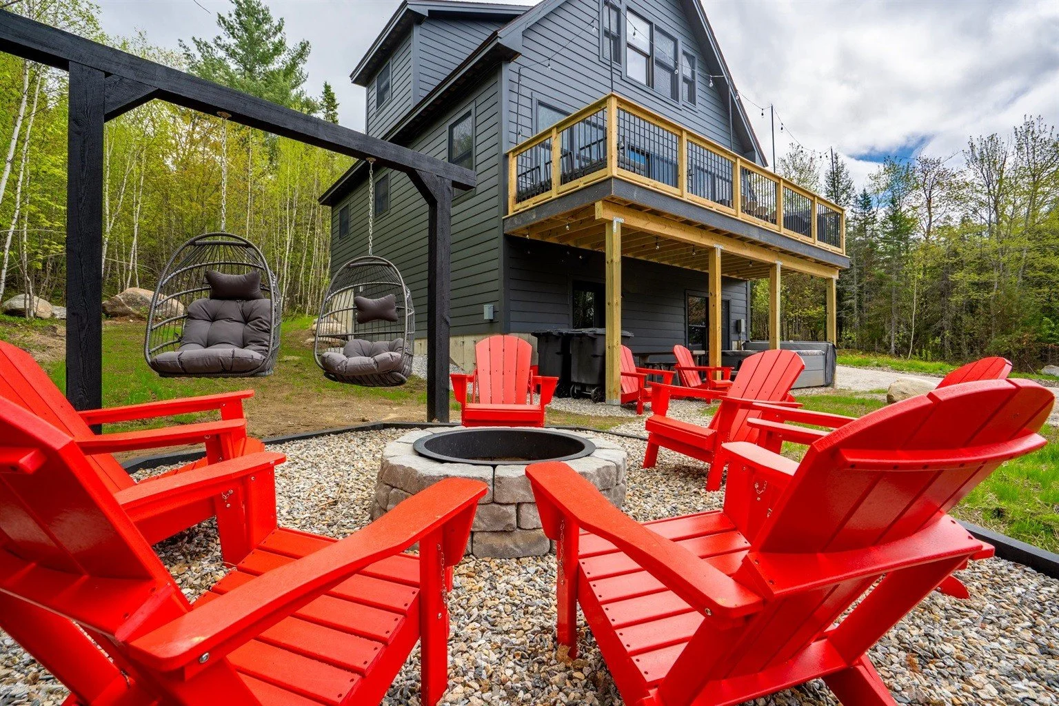 Outdoor seating area with red Adirondack chairs and a fire pit, surrounded by a house with a deck and hanging lounge chairs, in a wooded yard.