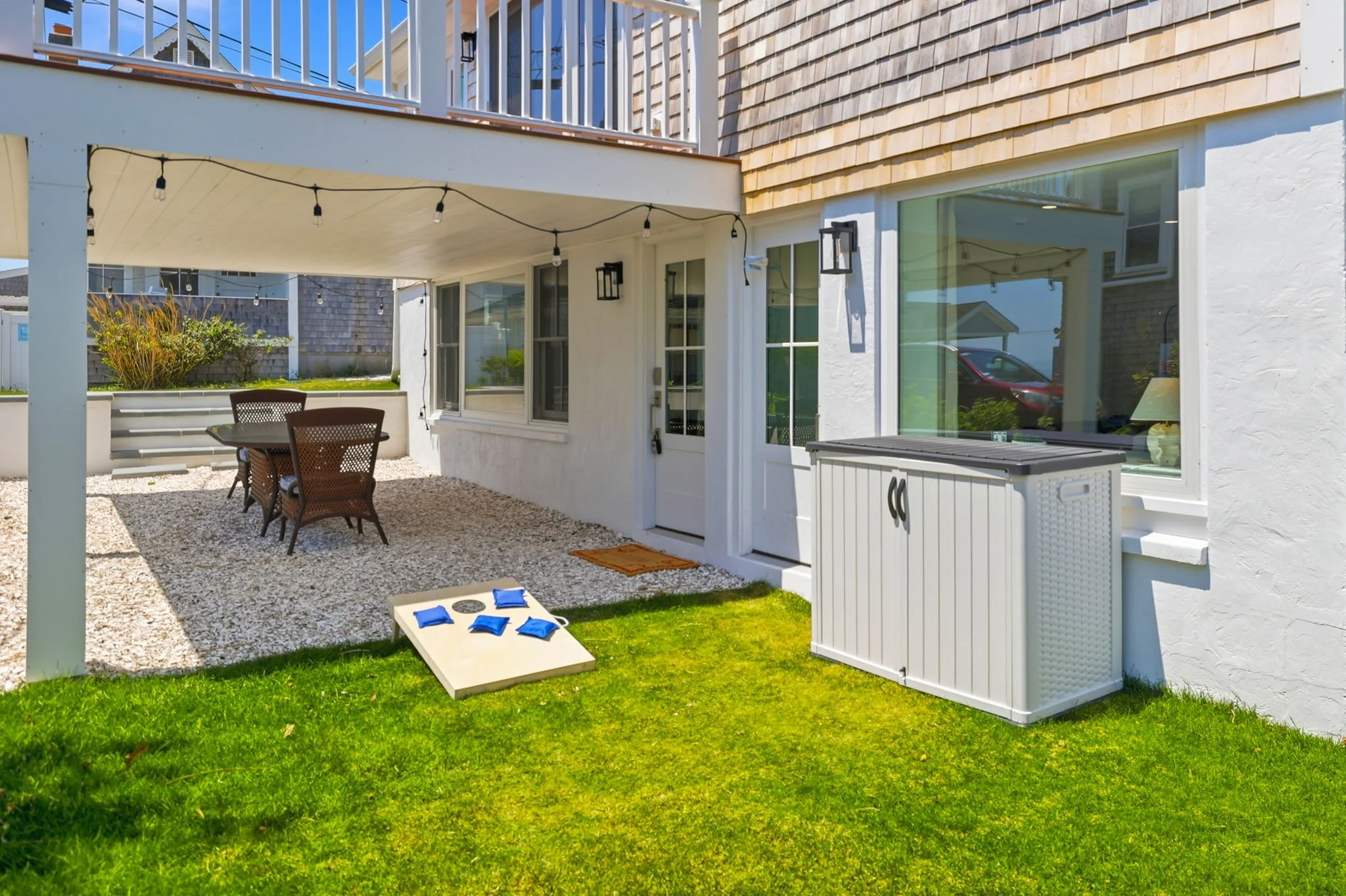 Backyard patio with patio table and chairs, string lights, small grassy area, window and door on house, shaded area with gravel, some outdoor storage, and a cornhole game on the grass.