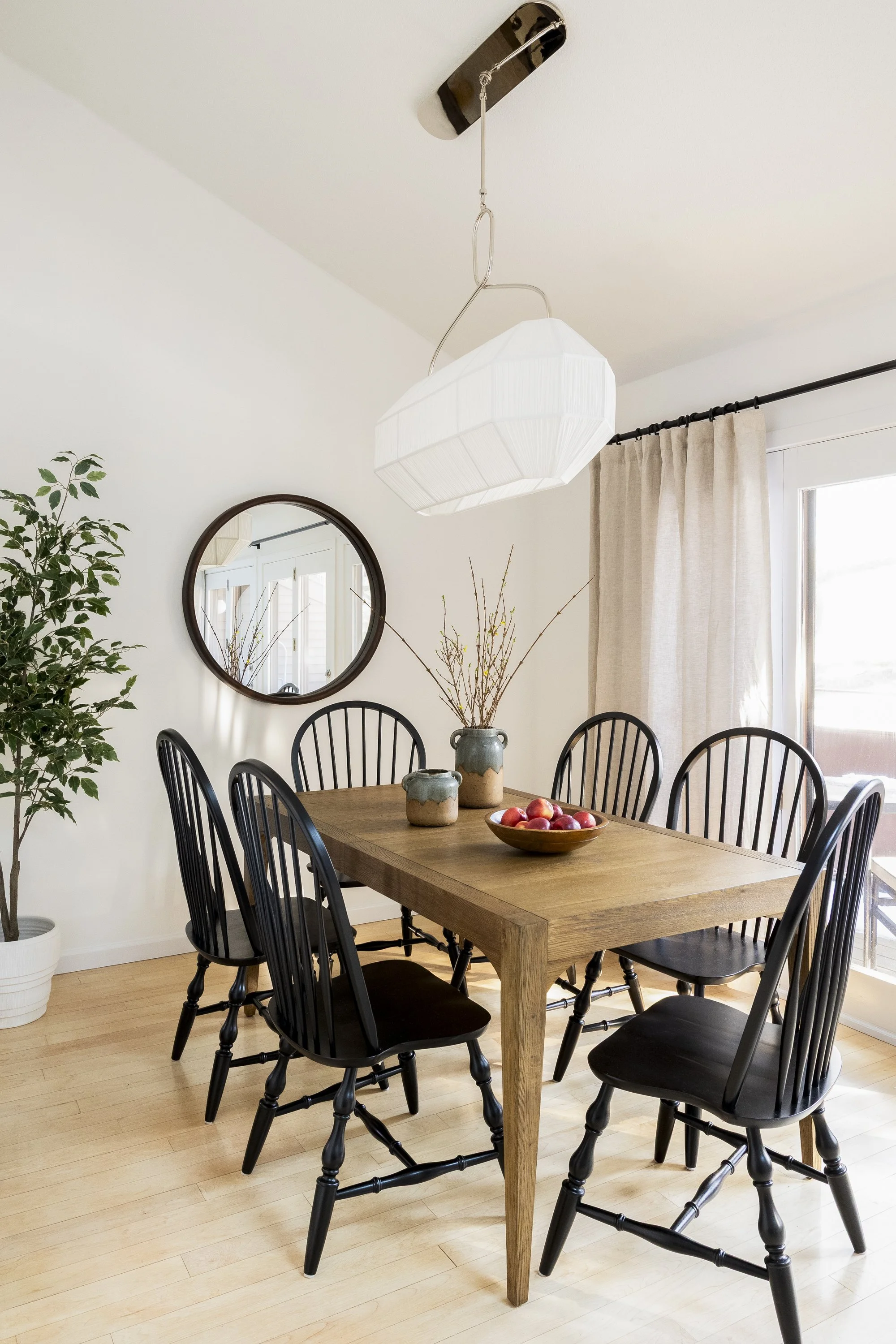 A wooden dining table with eight black chairs, a bowl of apples, three ceramic vases, a round mirror on the wall, a large window with beige curtains, a potted plant, and a white paper lantern pendant light hanging from the ceiling in a bright dining 