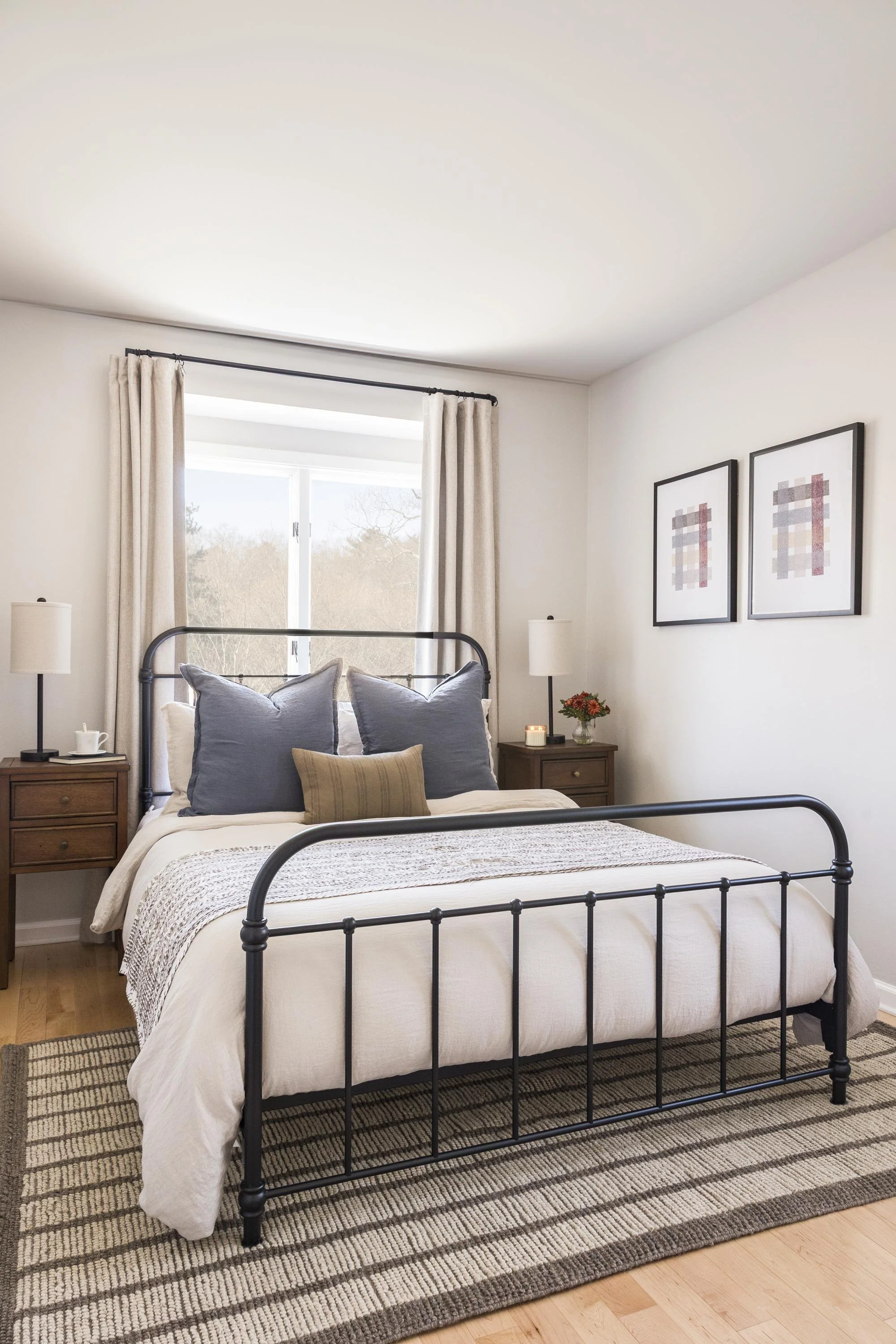 A bedroom with a black metal bed frame, white bedding, gray pillows, and a beige throw blanket. Two wooden nightstands with lamps and decor, a window with cream curtains, and framed artwork on the white walls.