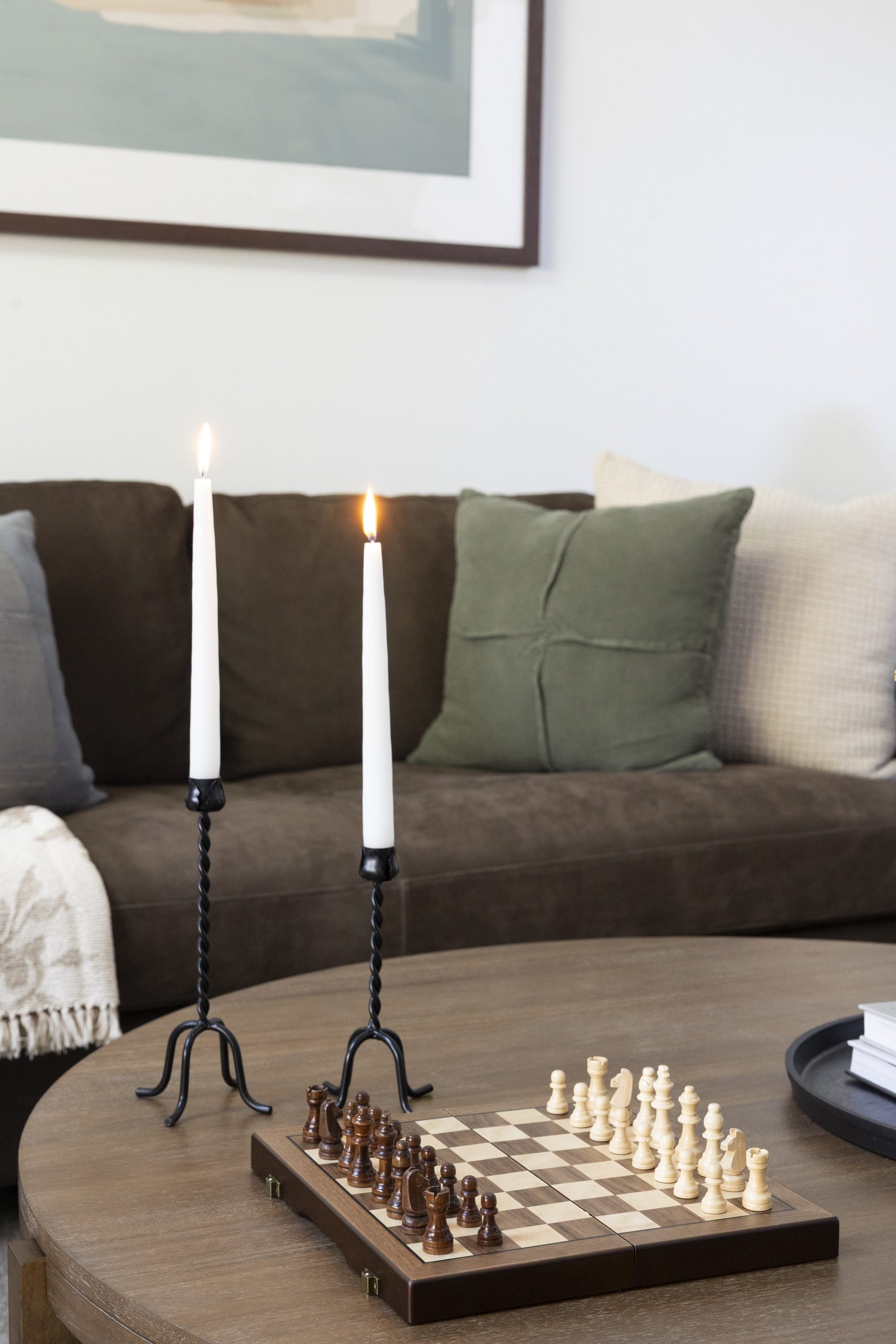 A chessboard set up for a game on a round wooden table, with a pair of lit white candles in black candle holders on the table, and a brown couch with green and beige pillows in the background.