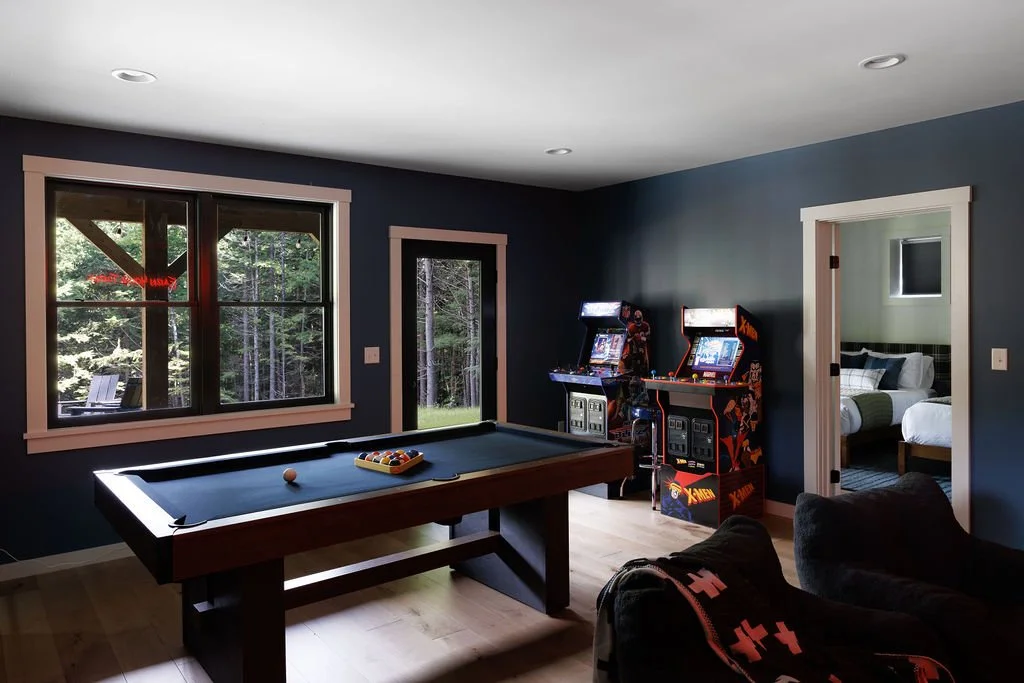 A game room with a pool table, two arcade gaming machines, a window showing trees outside, and a doorway leading to a bedroom with two beds.