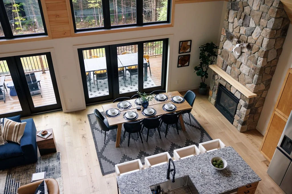 View of a spacious dining area with a wooden table set with plates and silverware, surrounded by black chairs, with ceiling-to-floor windows and a sliding glass door leading to an outdoor deck with a table and chairs, and a stone fireplace on the rig