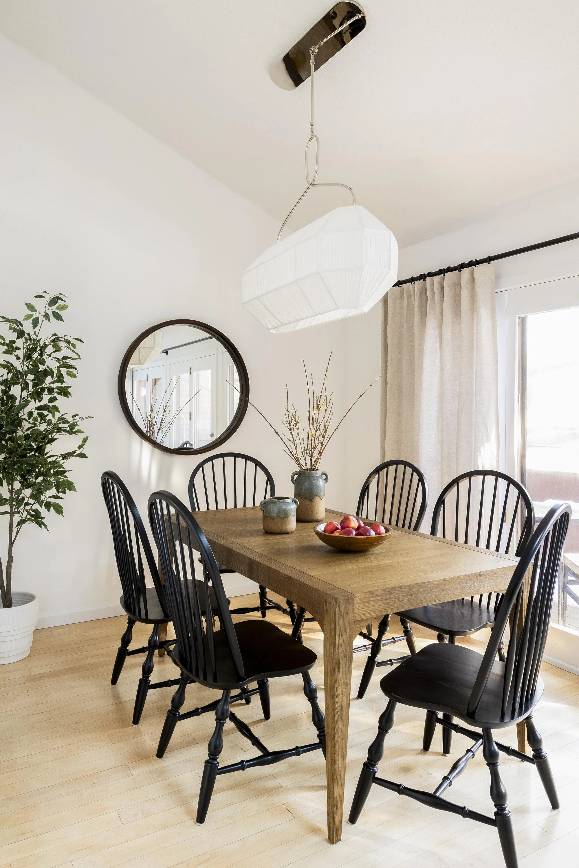 A dining room with a rustic wooden table surrounded by black Windsor-style chairs. A bowl of red apples is on the table, along with two ceramic vases. A round mirror hangs on the white wall, reflecting a door and window. A large white pendant light h