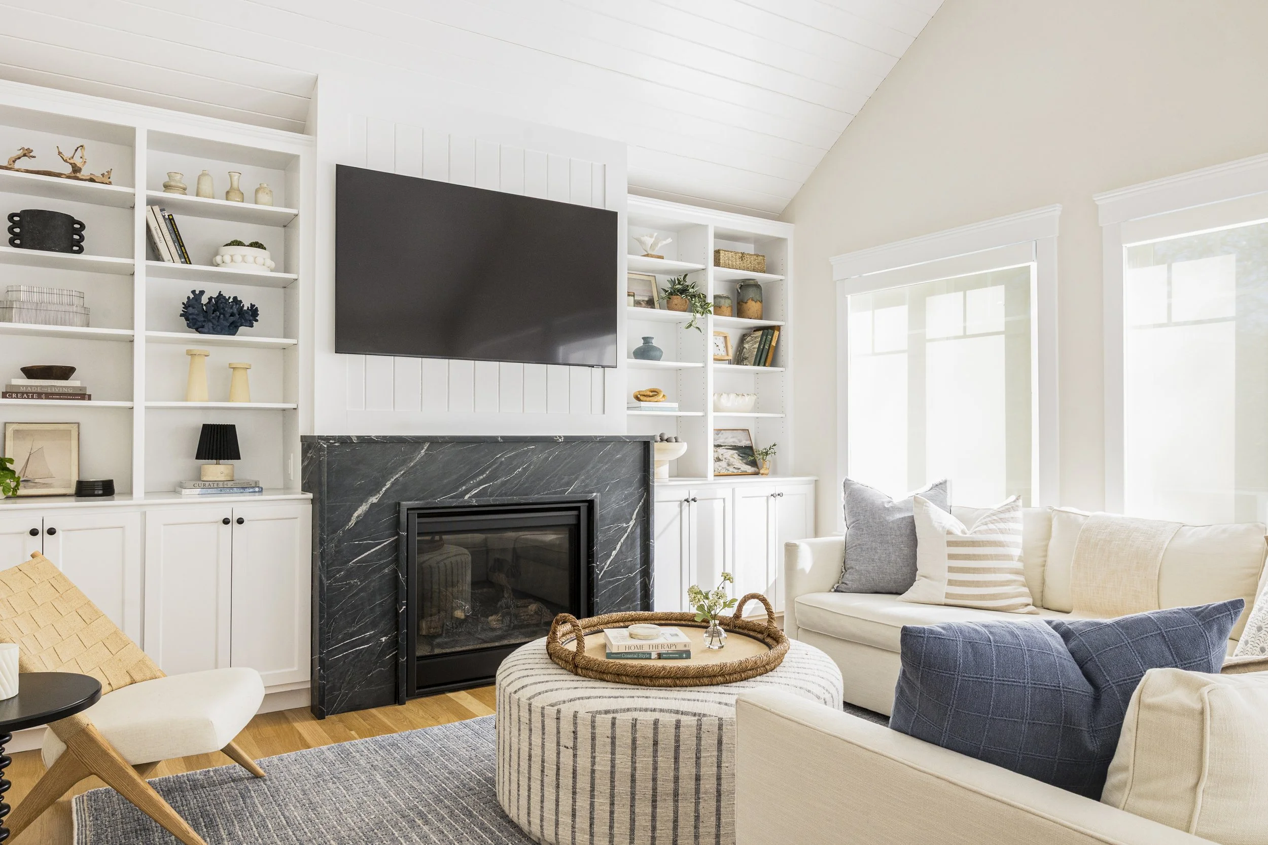 A cozy living room with a white sofa, a blue armchair, and a circular ottoman with books and a tray on it. A black marble fireplace with a large flat-screen TV above it, flanked by built-in white bookshelves filled with decorative objects and books. 