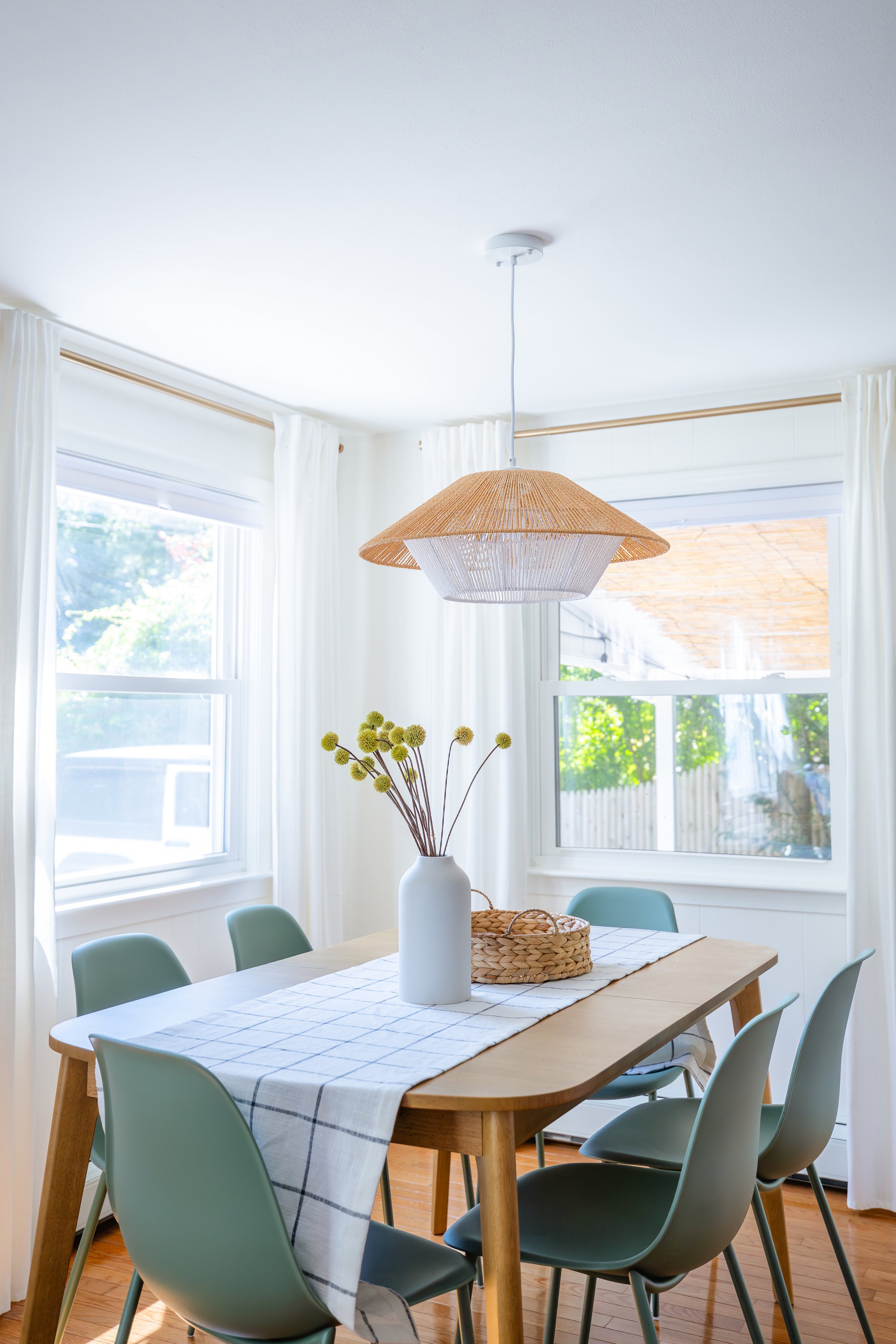 A dining room with a wooden table covered by a white and black checkered table runner. A white vase with yellow spherical flowers sits on the table. Light blue chairs surround the table. Large windows with white curtains let in natural light, and a v