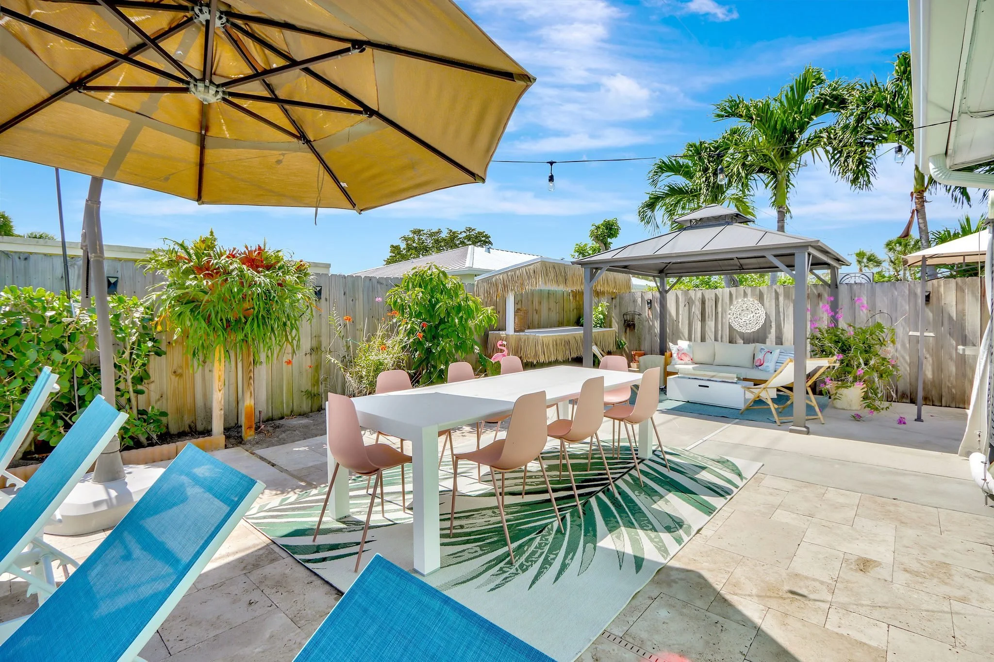 A backyard patio with outdoor furniture, including a white dining table with pink chairs, a white sofa with pink flamingo pillows, and beach chairs. There is a yellow patio umbrella, a thatched tiki bar, and lush green plants with a wooden fence and 
