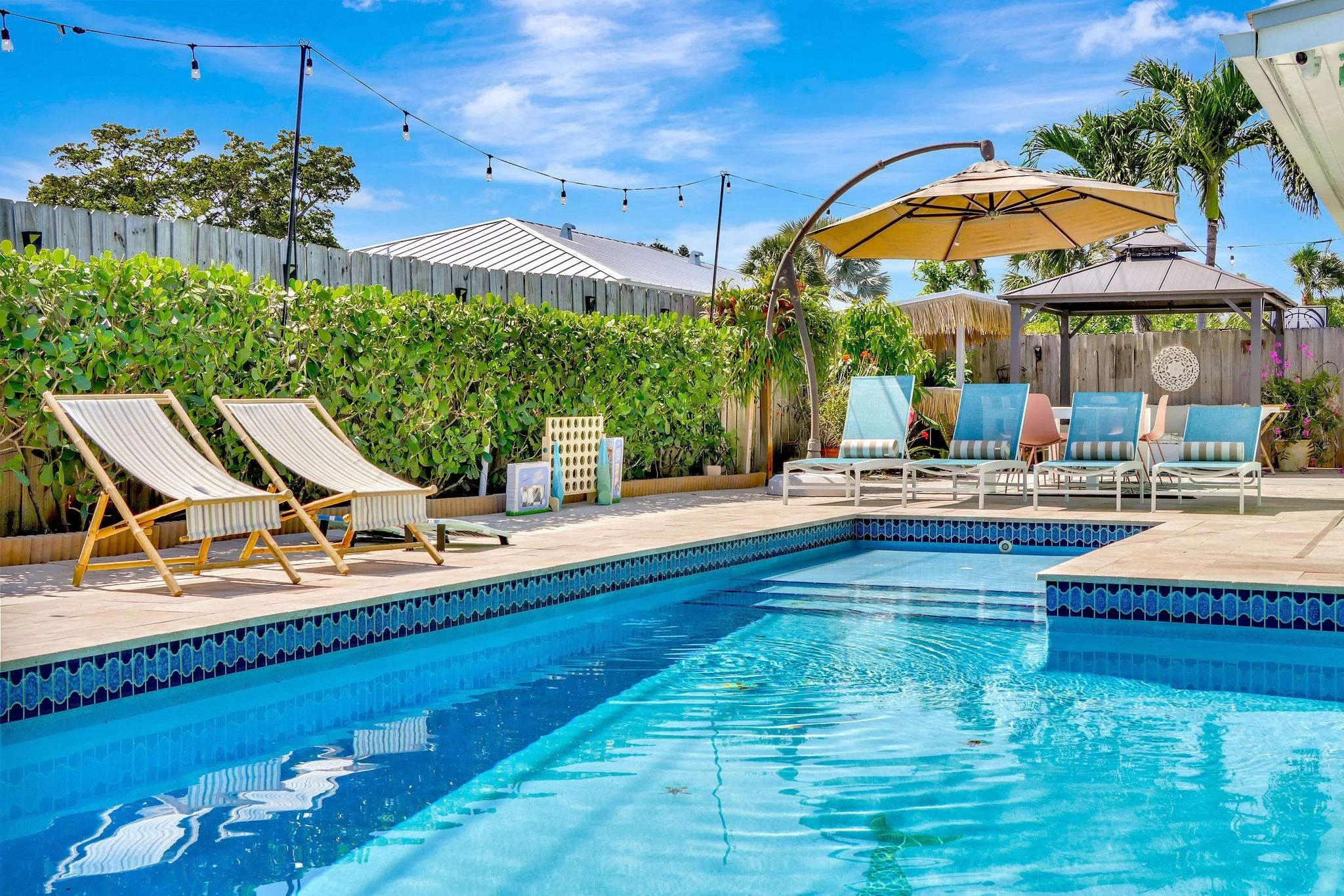 A backyard swimming pool with lounge chairs, umbrellas, and string lights under a blue sky.