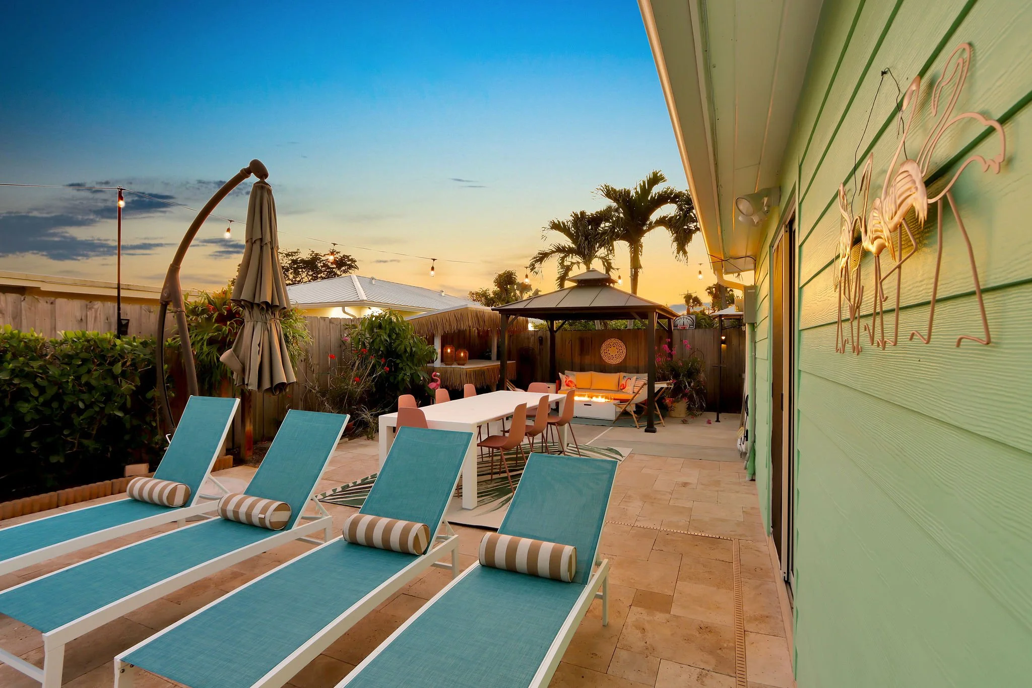 A backyard patio area with blue lounge chairs, a dining table with chairs, a green house wall with flamingo metal wall art, a gazebo, palm trees, string lights, and a sunset sky.