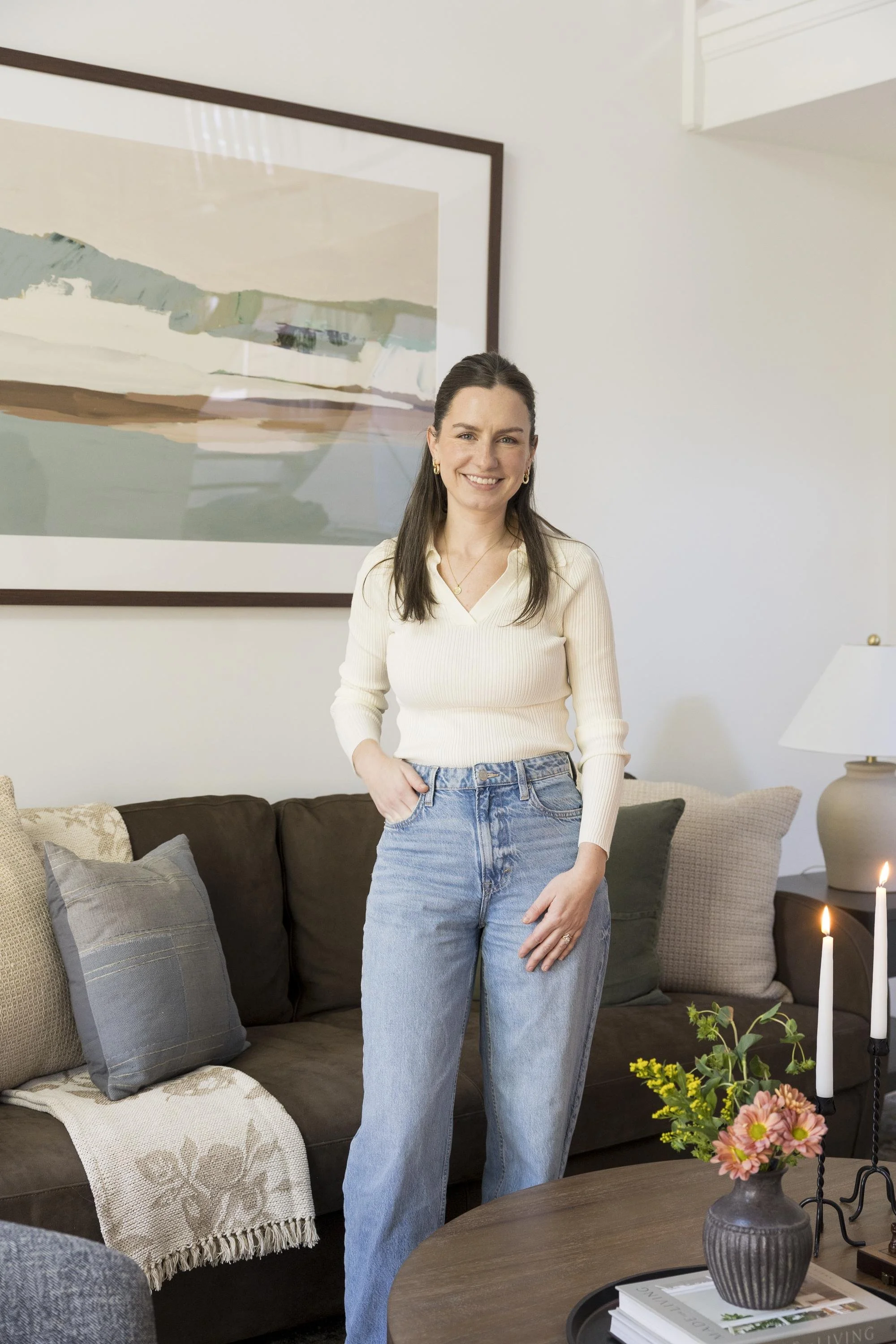 A woman stands in a living room, smiling at the camera. She is wearing a white long-sleeve top and blue jeans. The living room has a dark brown sofa with multiple decorative pillows, a gray and white throw blanket, a wooden coffee table with a vase o