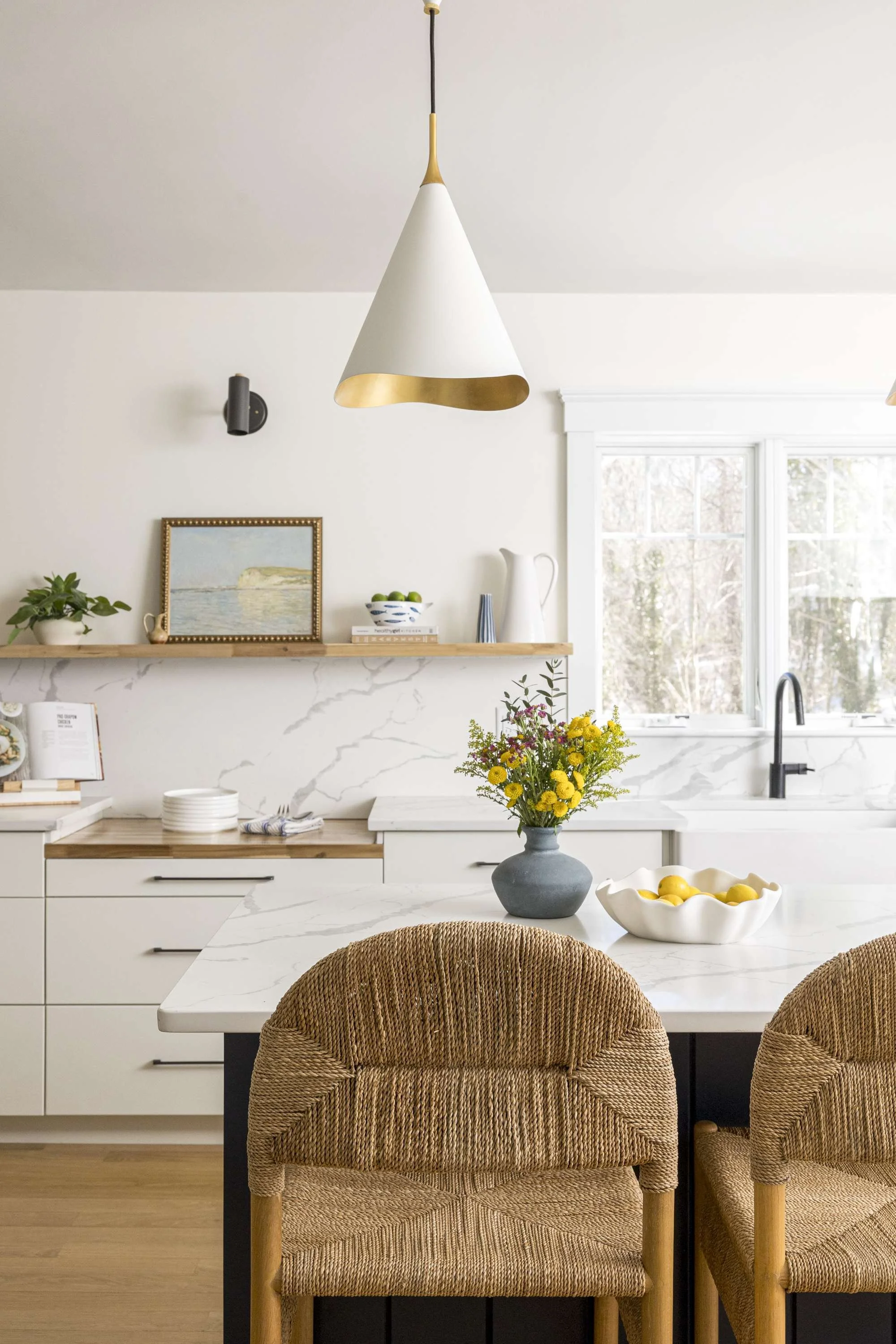 A modern kitchen with a marble countertop, a beige and gold pendant light, a window, and wicker chairs around a white marble table with a gray vase of yellow flowers.