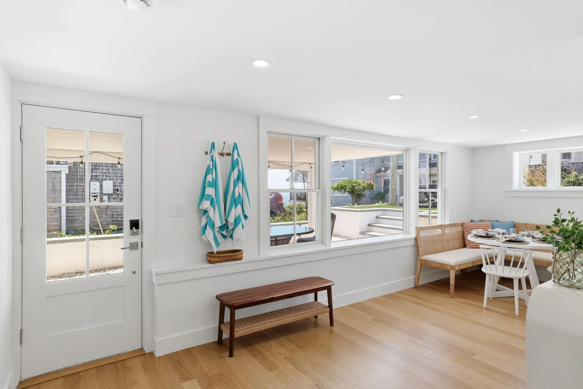 Bright sunlit indoor room with large windows, wooden floor, white walls, and a white door with glass panels. There are blue and white striped towels hanging on wall hooks, a wooden bench beneath the towels, and a small dining area with a white round 