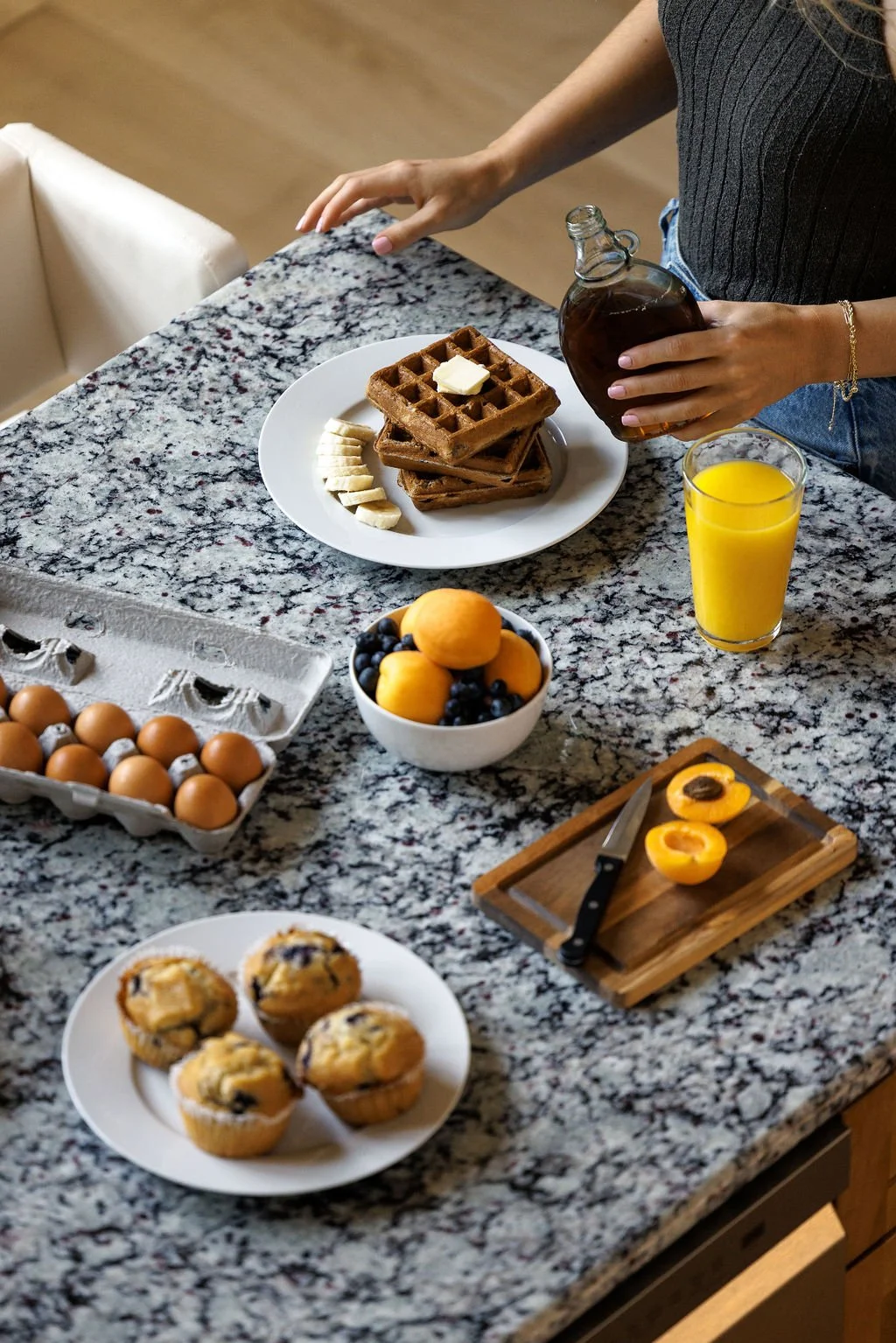 A woman preparing breakfast on a granite kitchen countertop, with waffles topped with butter, a banana, and blueberries, a carton of eggs, a bowl of mixed fruit, and a plate of muffins, along with a glass of orange juice and a bottle of syrup.