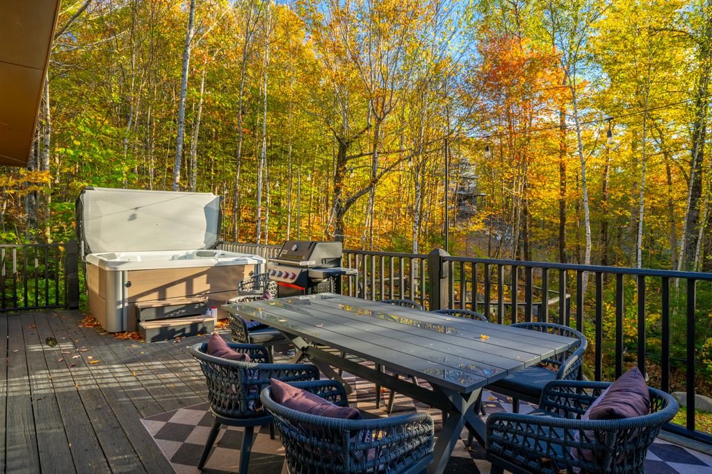 An outdoor wooden deck with a dining table, chairs, a grill, and a hot tub, surrounded by trees with fall foliage.