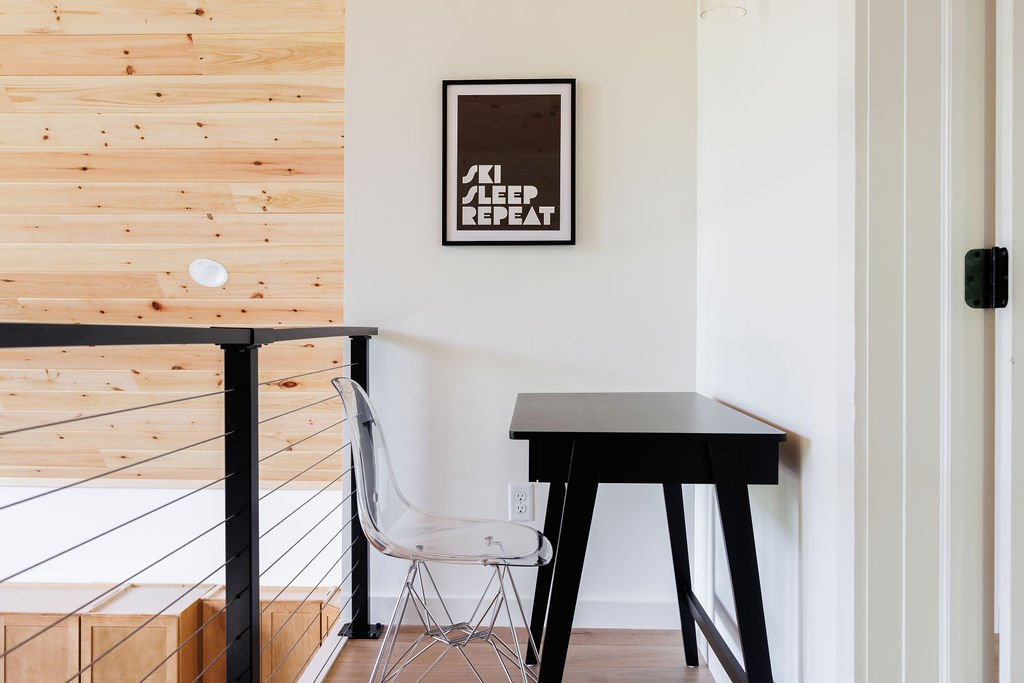 A corner of a room showing a black table, a clear plastic chair, a framed poster with the words "SKI SLEEP REPEAT" on a white wall, and a railing next to a wooden wall.