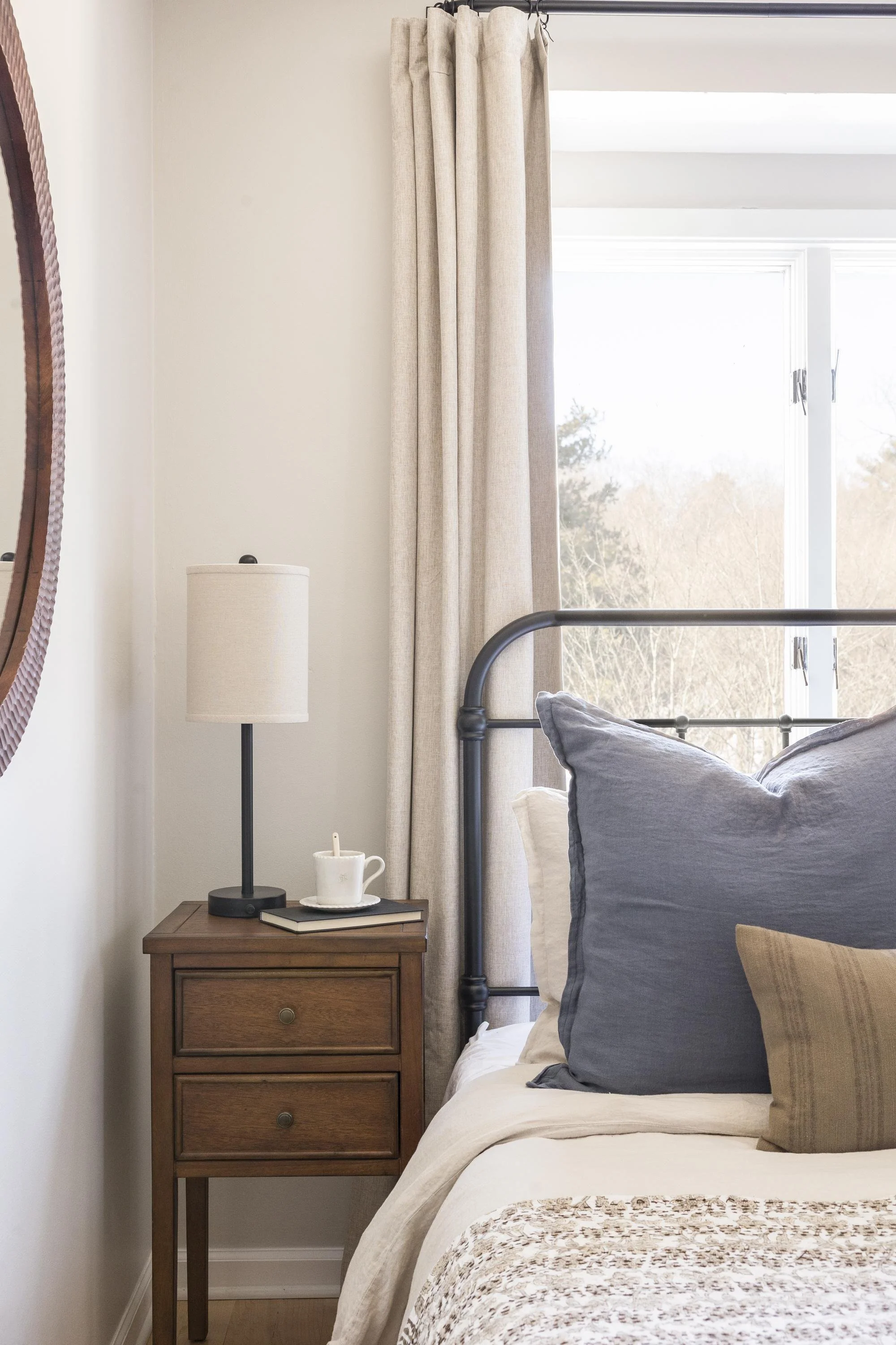 A bedroom corner featuring a wooden nightstand with a lamp, a Starbucks cup, and books. Next to the nightstand is a bed with a black metal headboard, decorated with pillows in gray, beige, and mustard tones. A window with beige curtains shows a view 