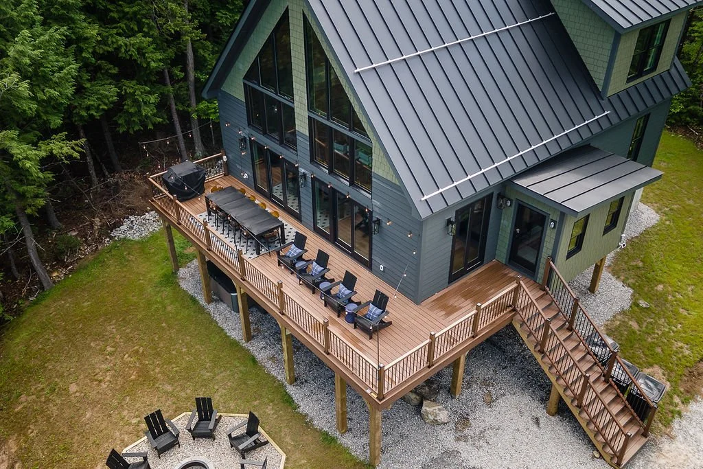 Aerial view of a modern house with a large wooden deck, outdoor furniture, and string lights, surrounded by trees and grass.