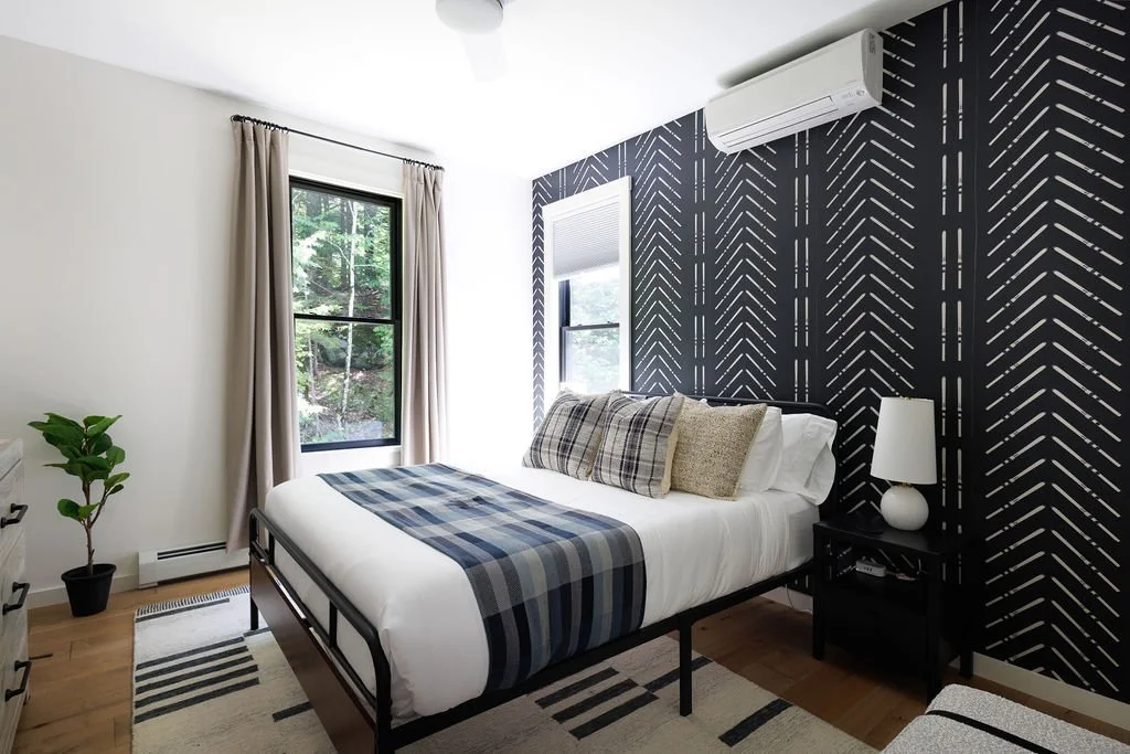 Bedroom with a bed, plaid blanket, pillows, side table with lamp, potted plant, windows with beige curtains, patterned accent wall, air conditioner, hardwood floor, and a rug.