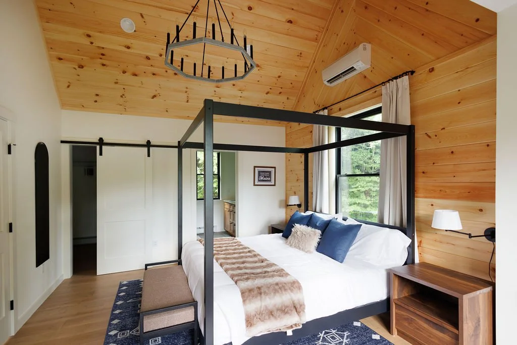 Bedroom with wooden ceiling and walls, black four-poster bed with white bedding and blue pillows, beige throw blanket and fur pillow, nightstand with lamp, large window with white curtains, air conditioning unit.