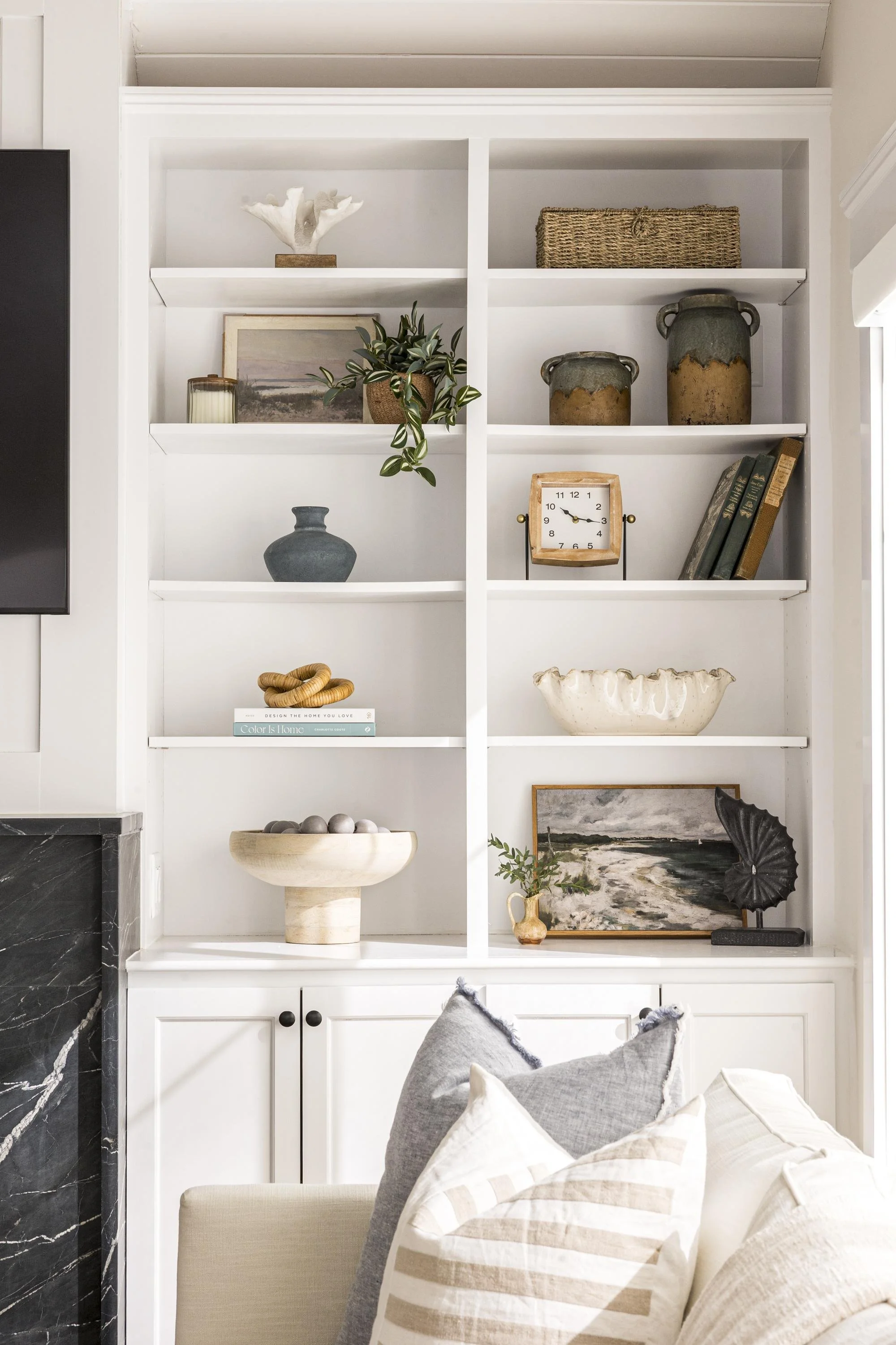 White built-in bookshelf with decorative objects, including ceramics, a plant, books, and framed art, in a living room with a beige sofa and pillows in the foreground.