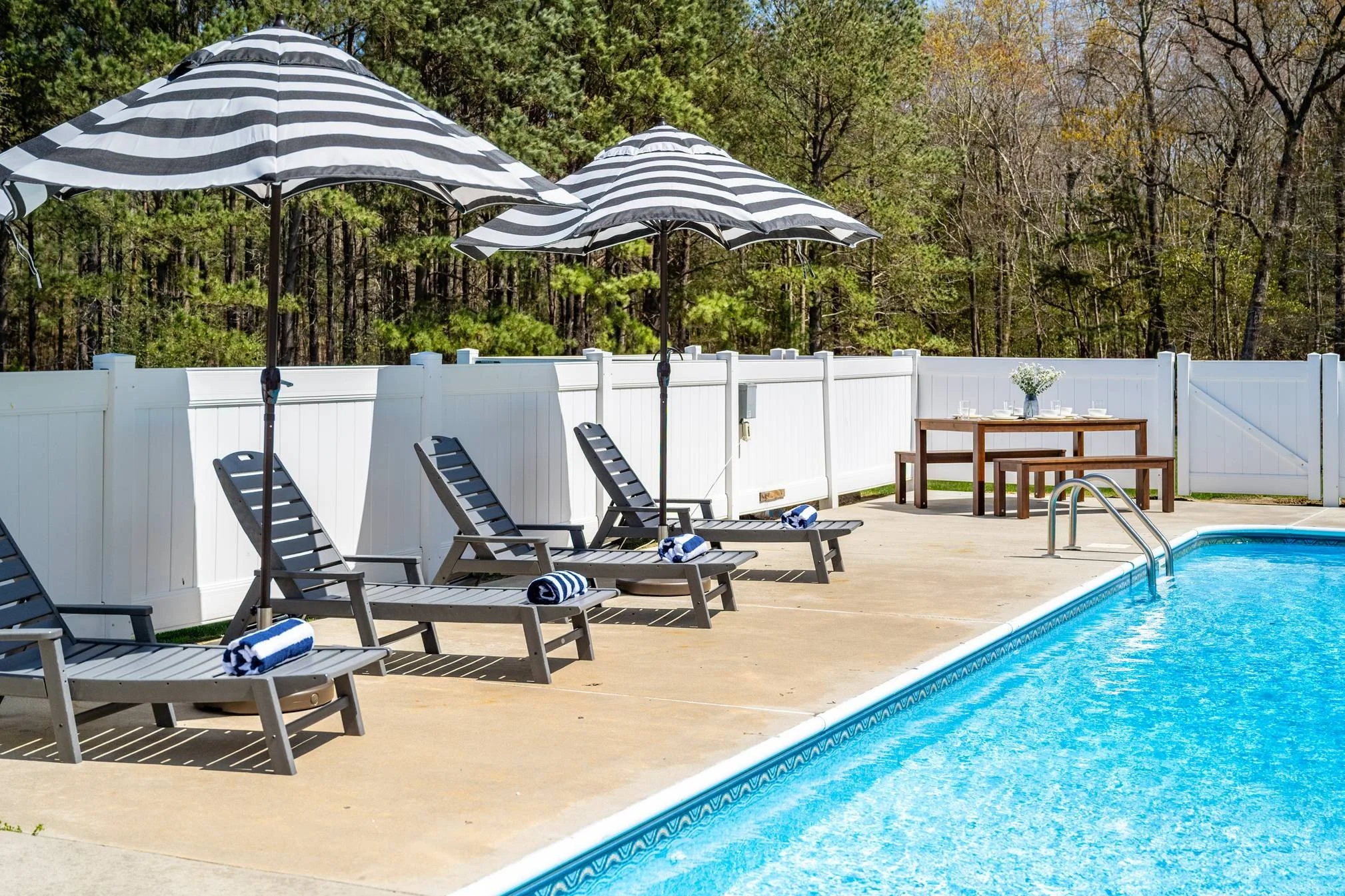 A backyard pool area with black and white striped umbrellas, lounge chairs with rolled towels, a table with a flower vase, and a white fence, surrounded by trees.