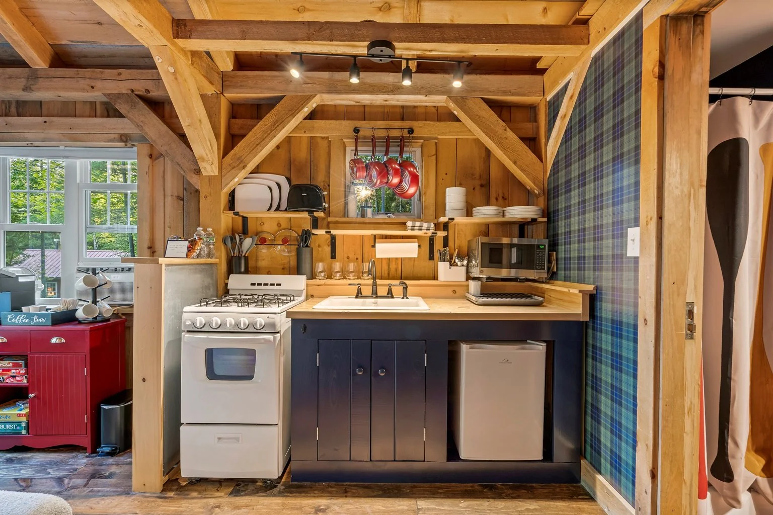 Compact kitchen with wooden walls and ceiling, featuring a white stove, small black cabinet with sink, microwave, mini fridge, and open shelving with dishes and utensils. Red hanging pots and pans are above the window, and a patterned blue-green wall
