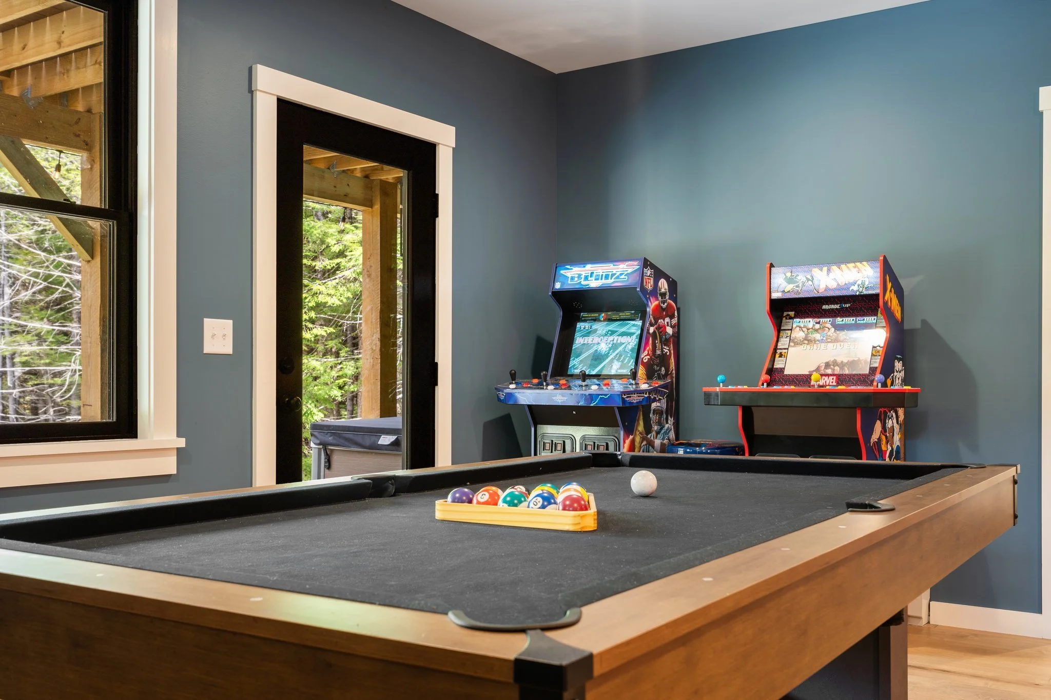 A pool table with a rack of billiard balls in the foreground. Two arcade game machines, one NFL Blitz and one X-Men, are against a blue wall in a recreational room. A door and a window showing a wooded outdoor area are in the background.