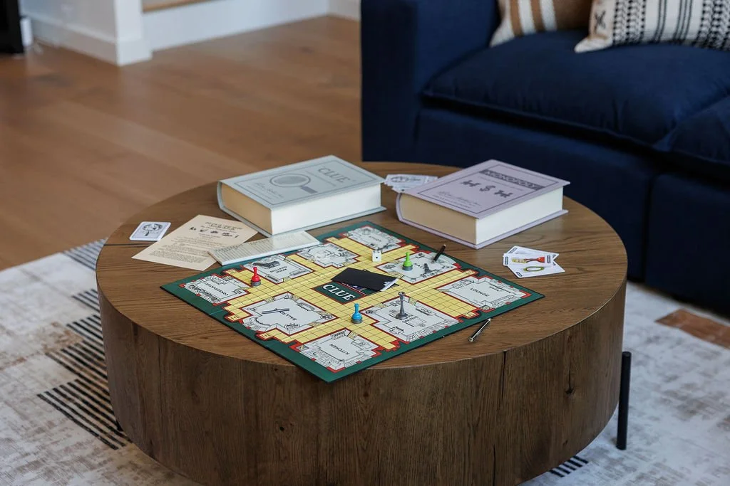 A board game set up on a round wooden coffee table in a living room with a navy blue couch. The game includes a game board with colorful tracks, miniature game pieces, and playing cards, along with some instruction booklets and papers nearby.