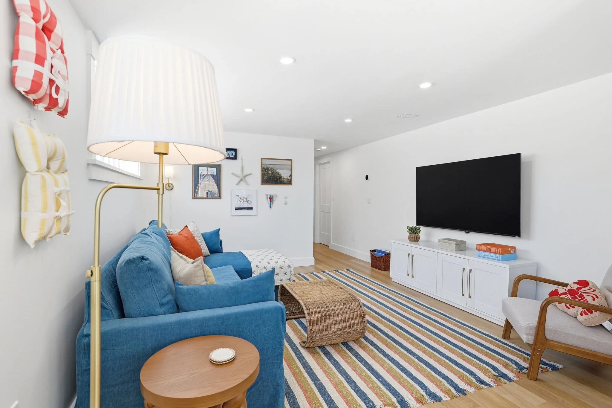 A cozy living room with a blue sofa, yellow and orange pillows, a small wooden side table, a woven ottoman, a colorful striped rug, a white TV console with books, a potted plant, wall art, and a white wall with decorative pillows on the left
