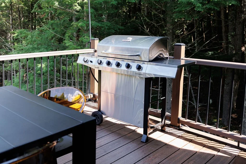 A stainless steel outdoor grill on a wooden deck next to a glass table with a yellow-tinted umbrella, surrounded by a black metal railing with trees in the background.