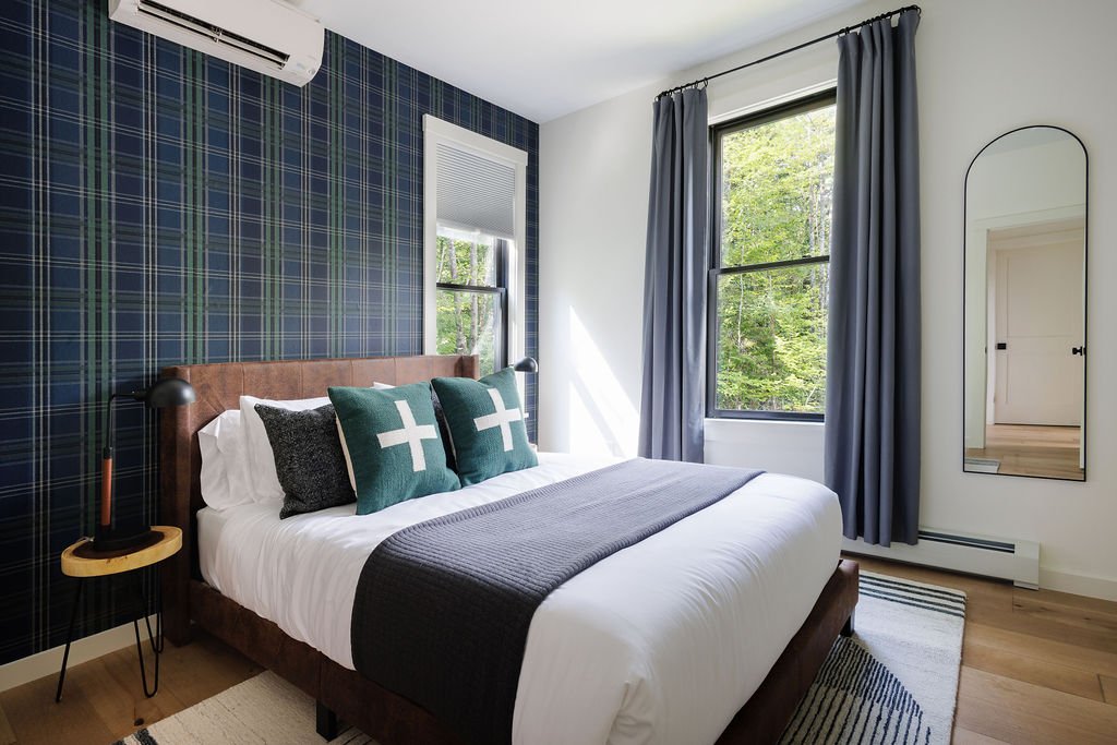 A bedroom with a bed, dark plaid accent wall, two pillows with white crosses, and a window showing greenery outside.