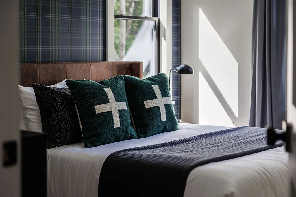 A neatly made bed with dark green pillows featuring white cross designs, a brown headboard, and a black blanket, in a bright bedroom with a window and plaid wallpaper.