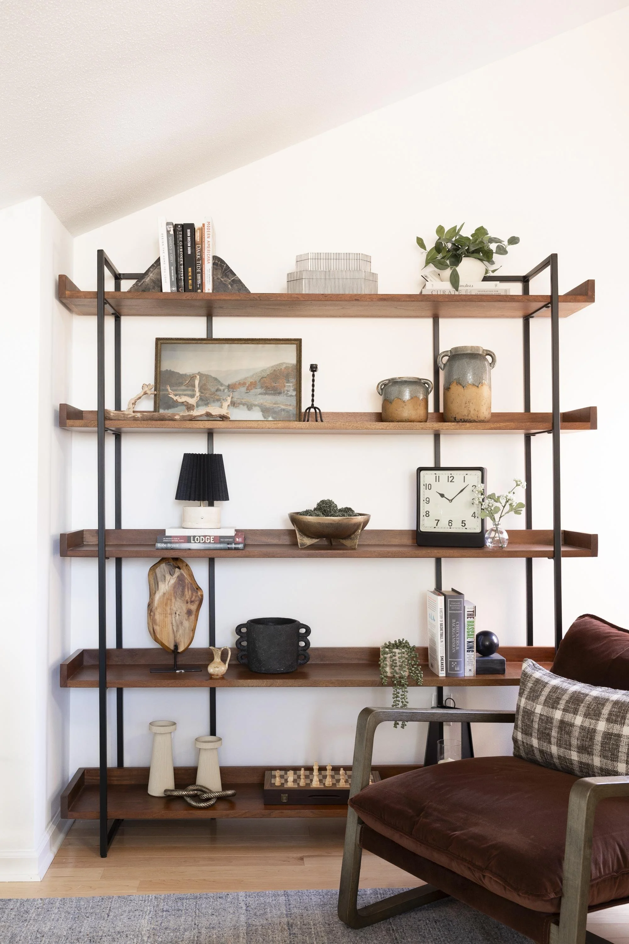 A wooden bookshelf with black metal frame decorated with books, pottery, artwork, and plants, situated next to a brown chair with plaid pillow on a light wood floor.