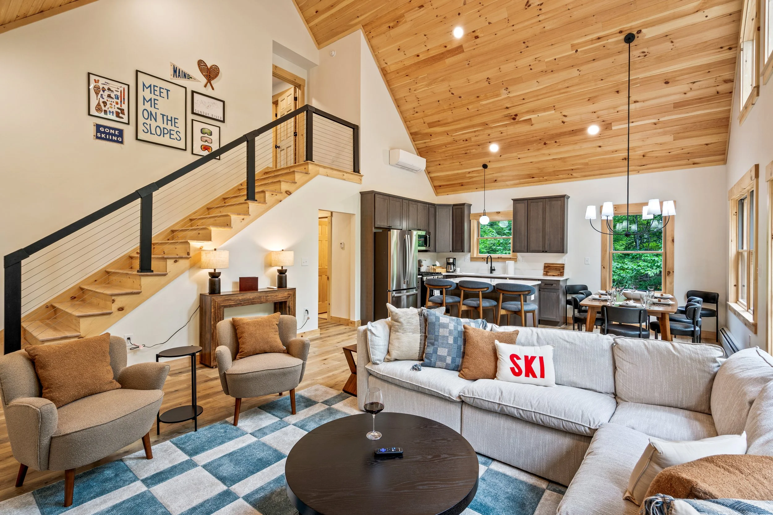 Open-concept living room and kitchen with wooden vaulted ceiling, beige couch with pillows, armchair, coffee table with remote and wine glass, kitchen with dark cabinets, stainless steel appliances, bar stools, and large windows showing green trees outside.