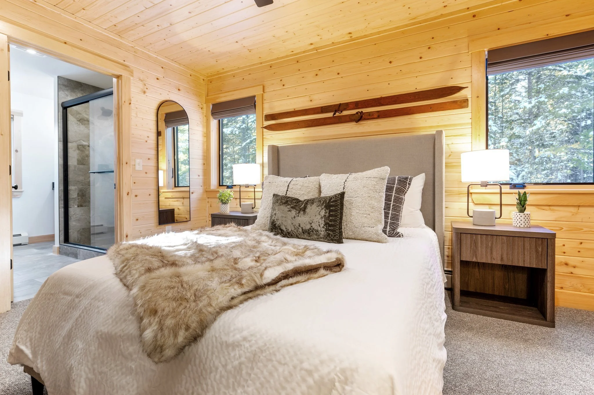 A cozy bedroom with wood-paneled walls and ceiling, featuring a bed with cream-colored quilt, decorative pillows, faux fur throw, two nightstands with lamps, a large window showing trees outside, and a view of a bathroom with a glass shower door.