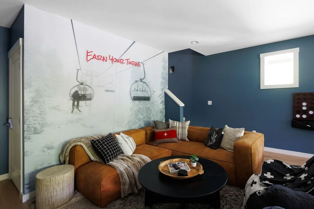 Living room with a tan sectional sofa, throw pillows, a black coffee table, and a wall mural of snow-covered trees and a ski lift with the phrase "EARN YOUR TURNS" written in red.