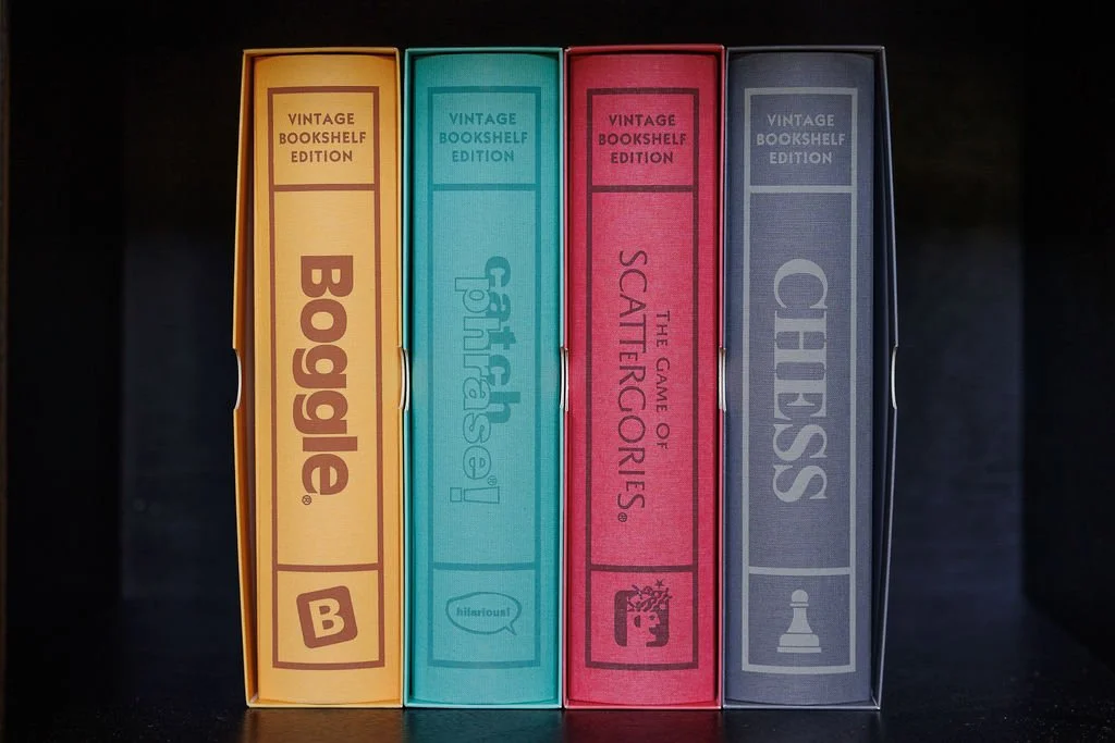Four vintage board game boxes are standing upright on a black surface. From left to right, they are titled 'Boggle,' 'Cattails,' 'The Game of Scattergories,' and 'Chess.'