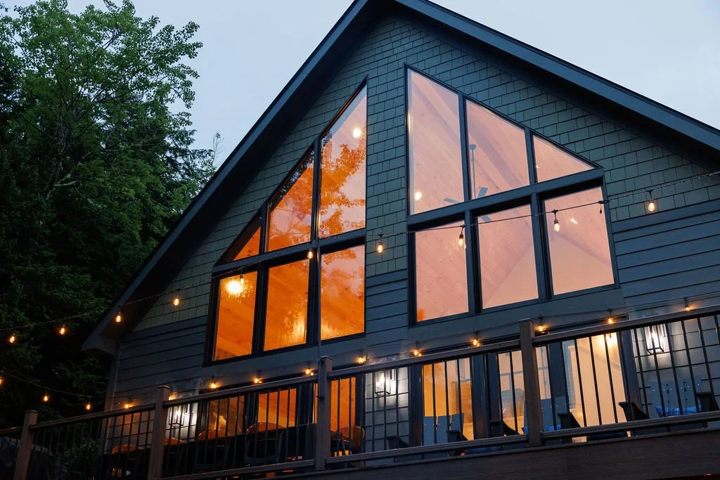 A modern house with large triangular windows on the upper floor, surrounded by string lights and trees outside at dusk.