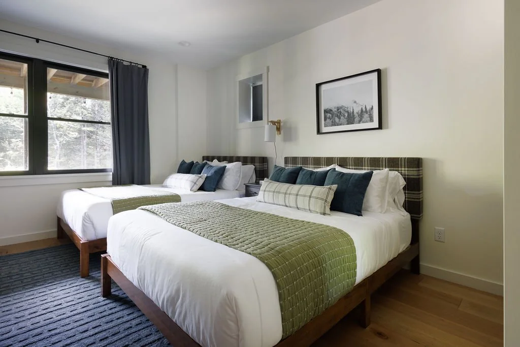 Two neatly made beds in a bright, modern bedroom with forest-themed artwork and a large window with dark curtains.