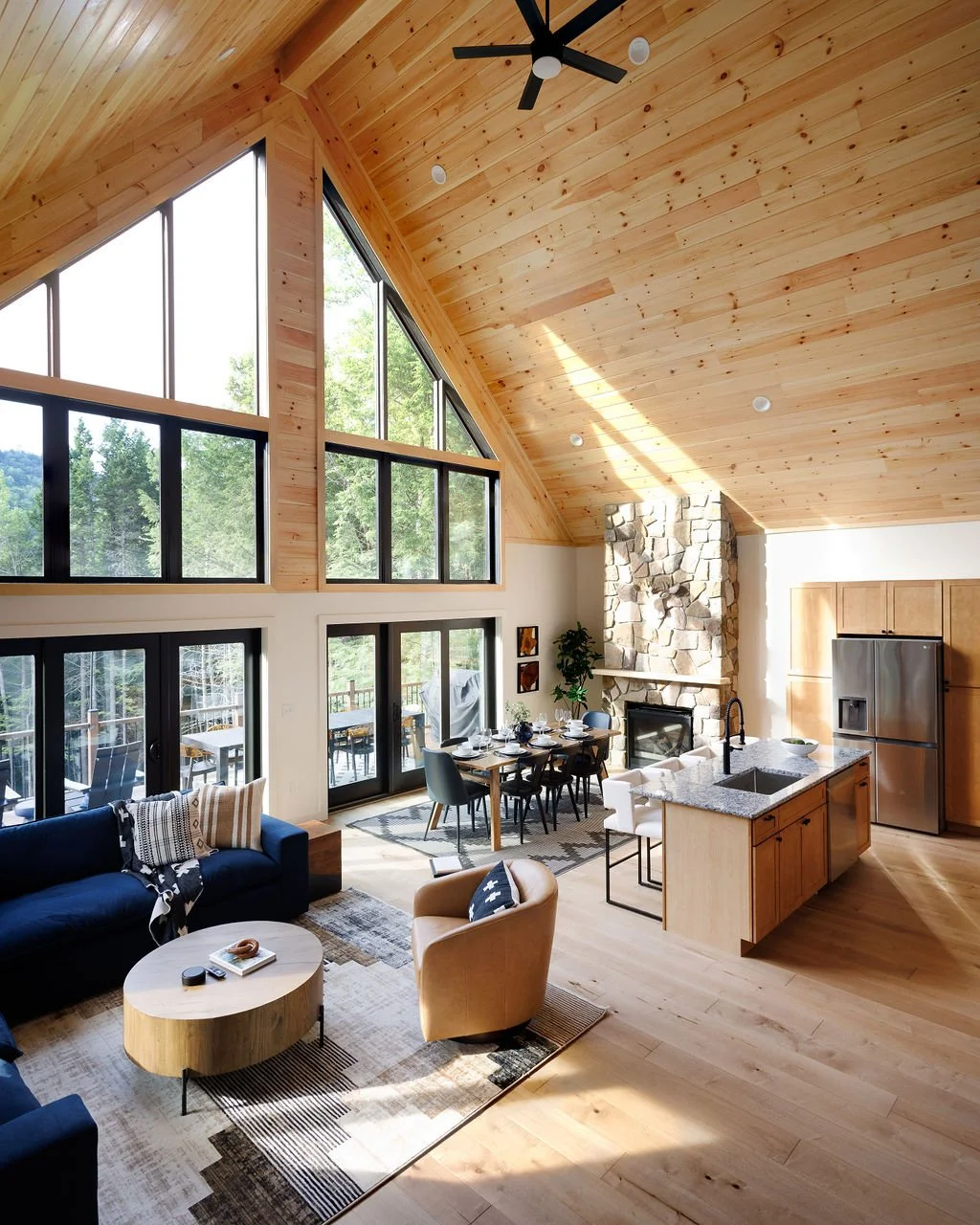 Open-concept living room and kitchen with vaulted wood ceiling, large windows, stone fireplace, dining table, sofa, armchairs, and kitchen island.